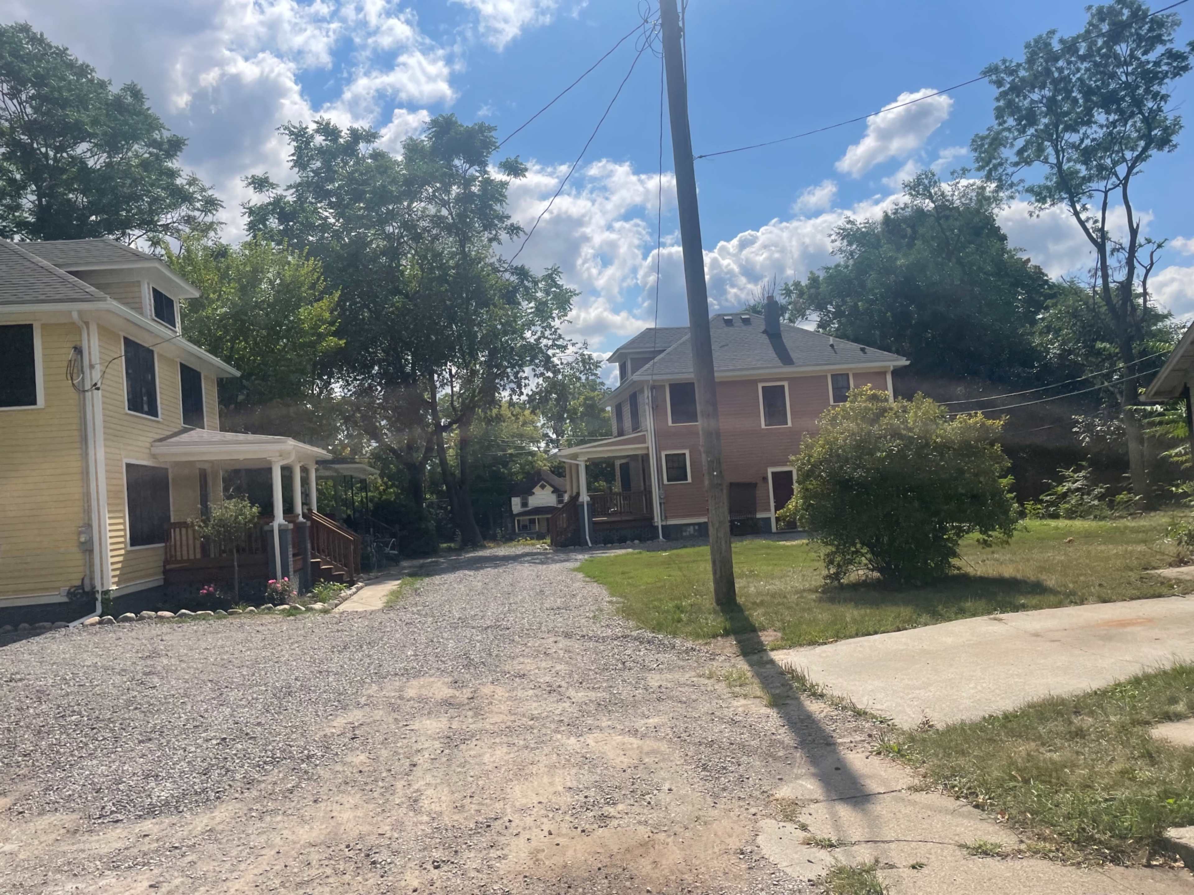 Two houses line a gravel driveway under a partly cloudy sky, with power lines overhead and trees in the background.