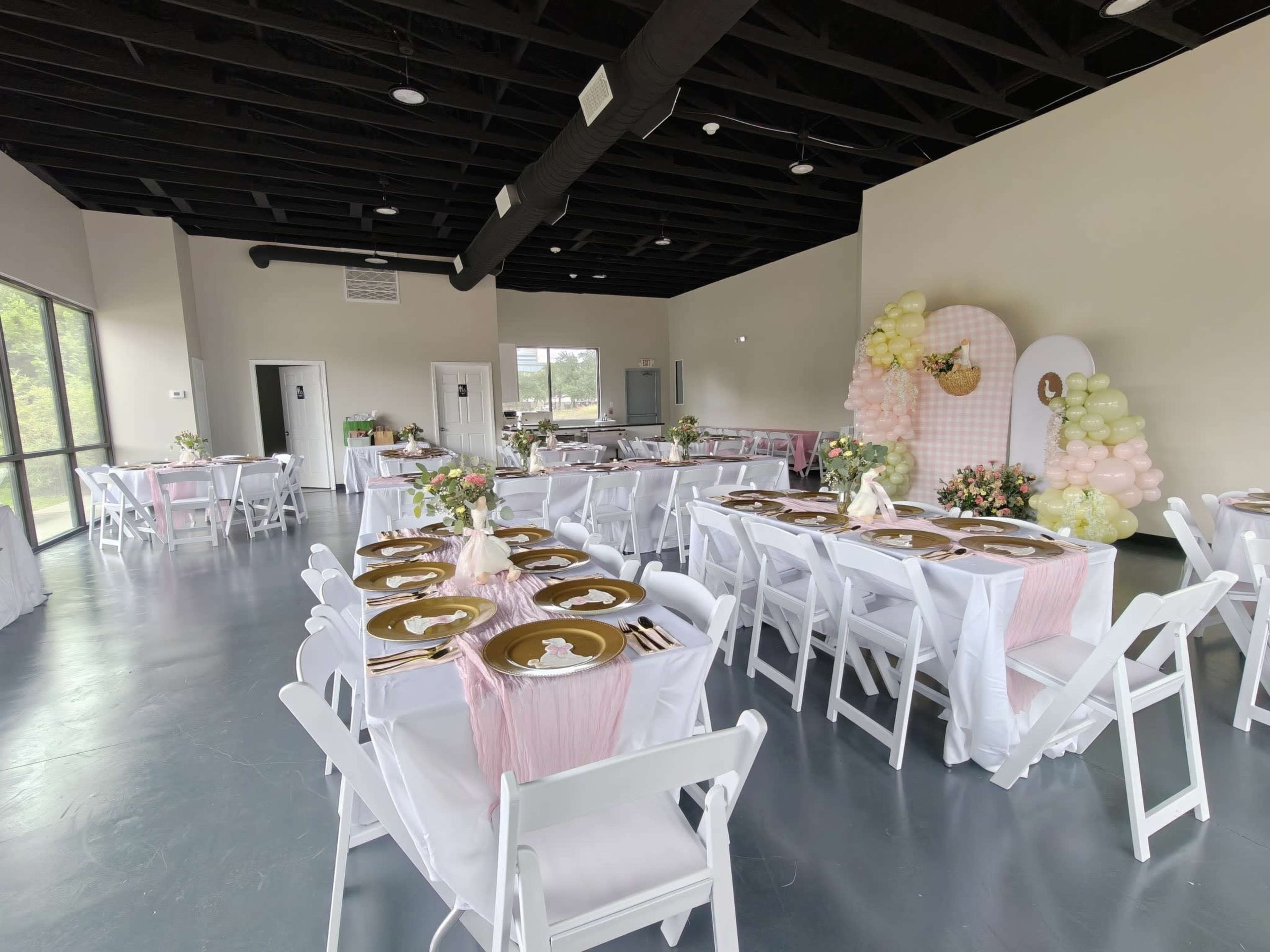The image shows a spacious event venue set up for a celebration, featuring tables adorned with white tablecloths, gold chargers, floral centerpieces, and a backdrop decorated with pastel balloons.