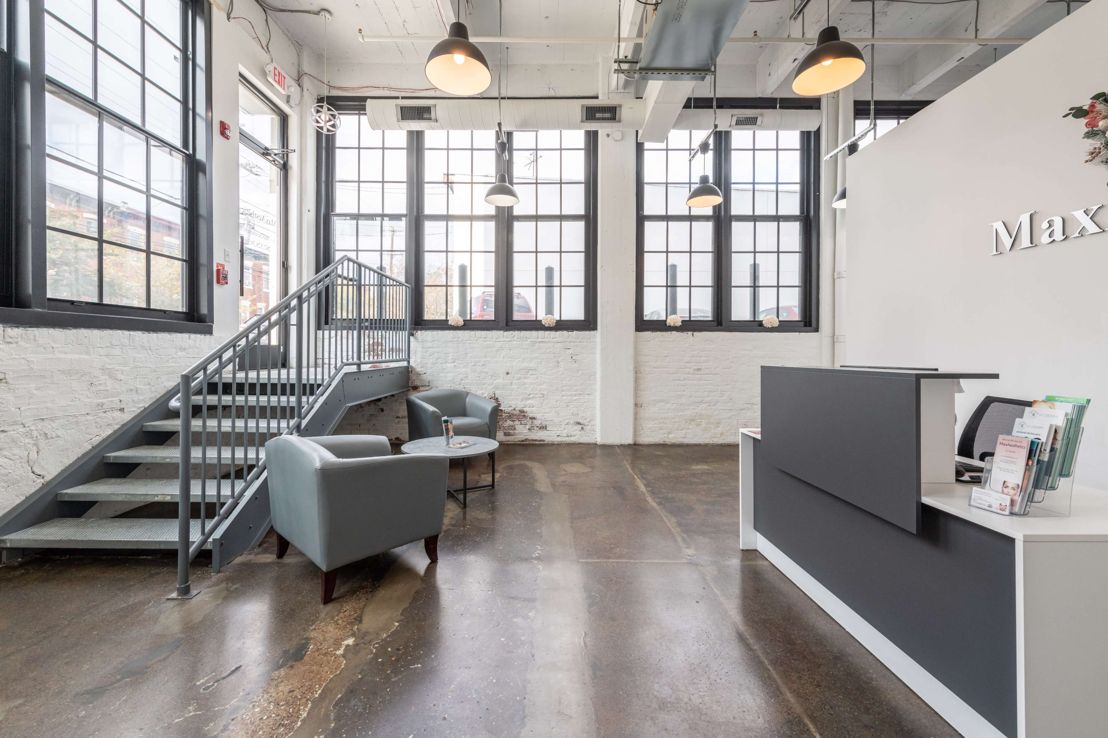 The image shows a modern reception area with large windows, a small seating area with two chairs, and a staircase leading to an upper level.