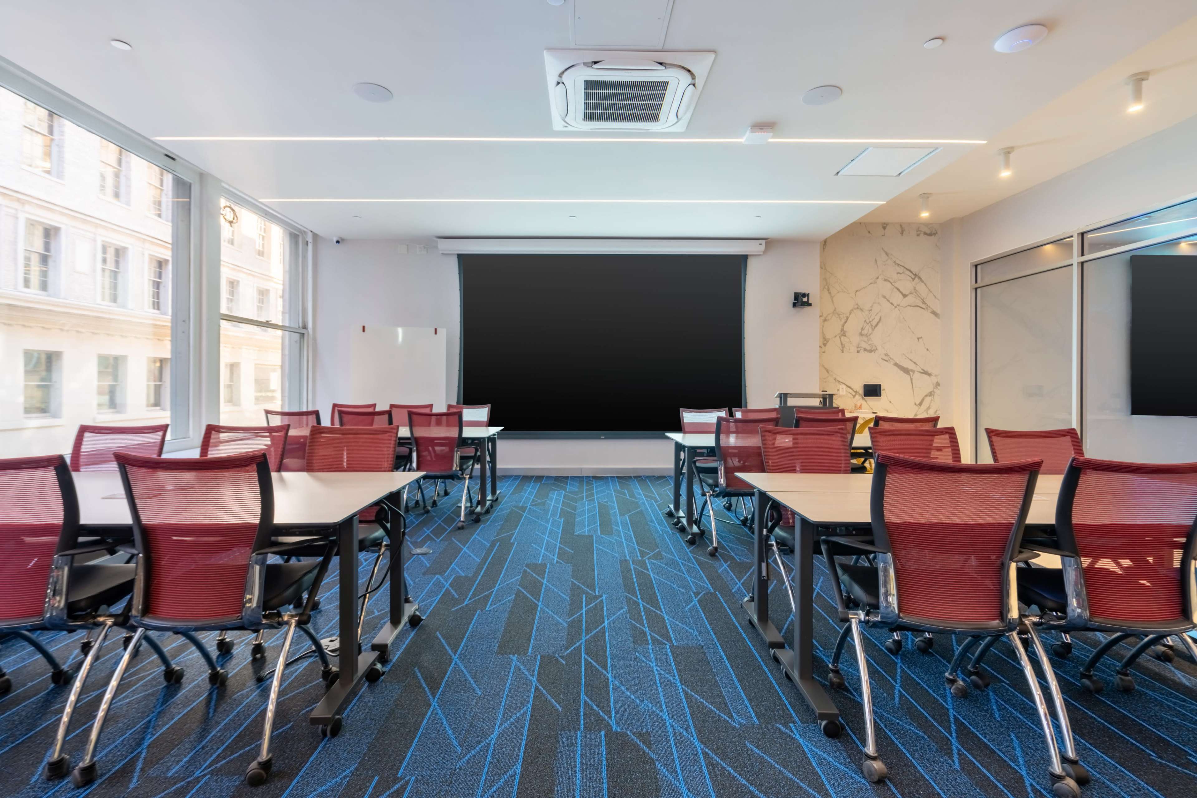 A modern conference room features rows of red chairs facing a large screen and large windows allowing natural light.