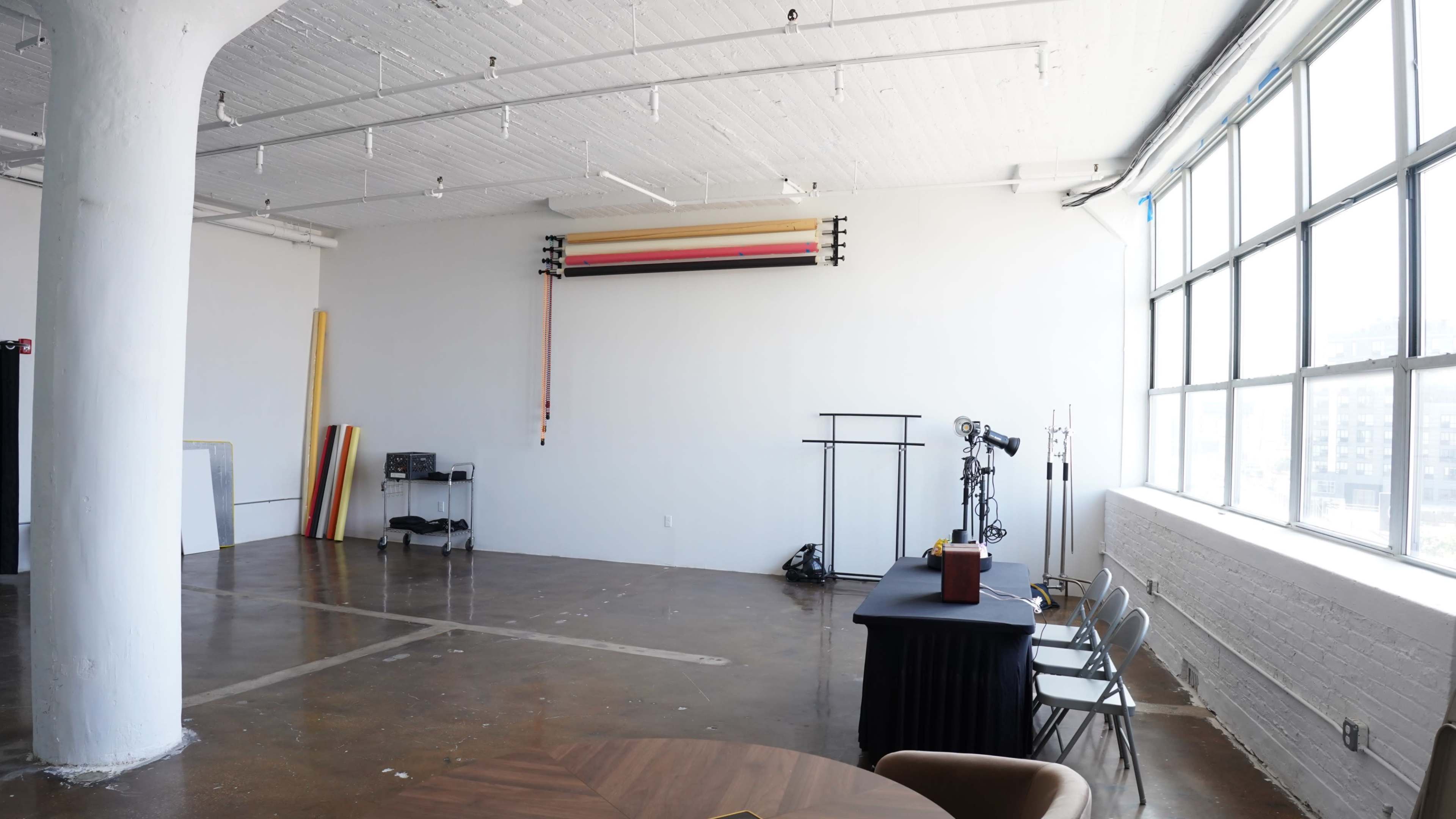 The image shows a spacious, empty studio with large windows, a plain white wall featuring rolled backdrops, and a table with chairs set up in the corner.