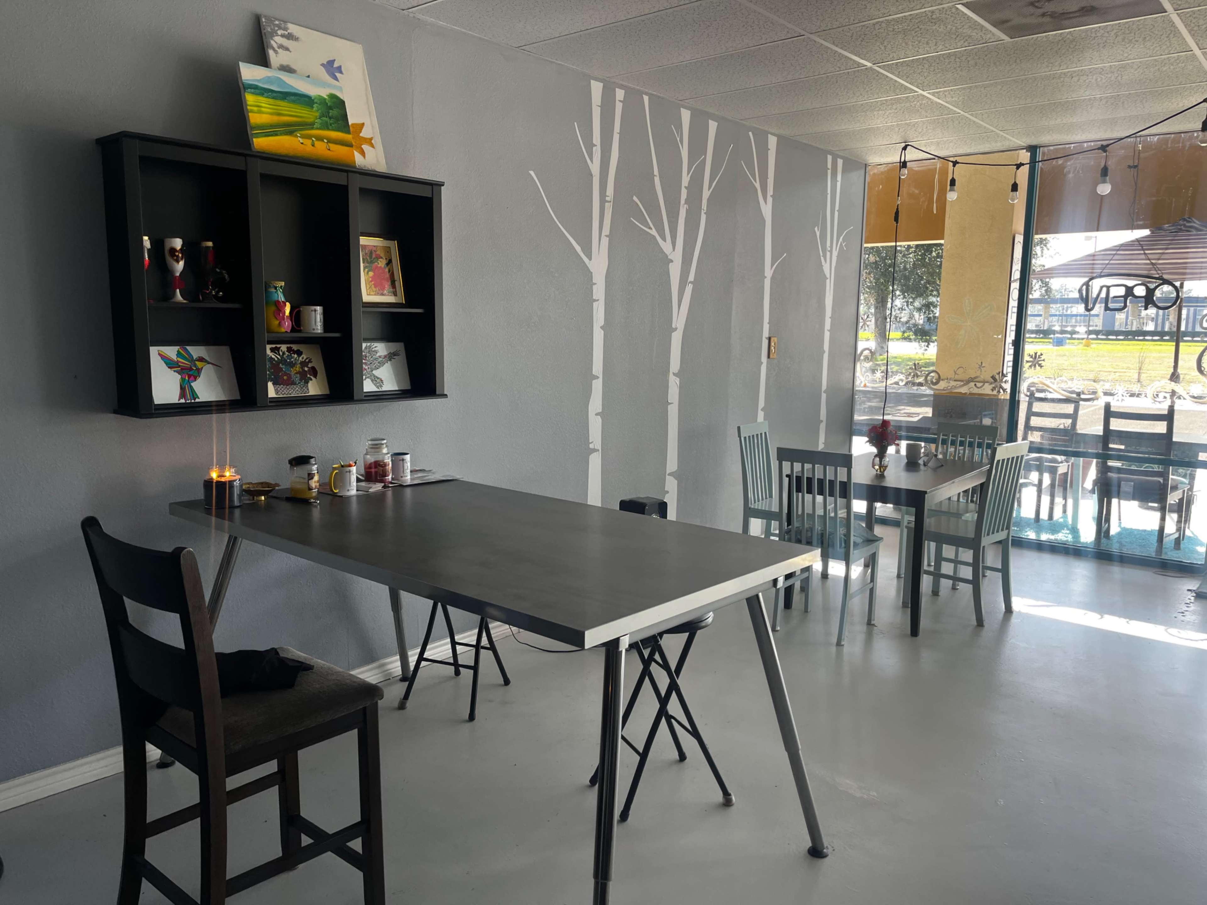 A minimalist café interior featuring a black table with folding legs, a shelf with decorative items, and white tree murals on the gray walls.