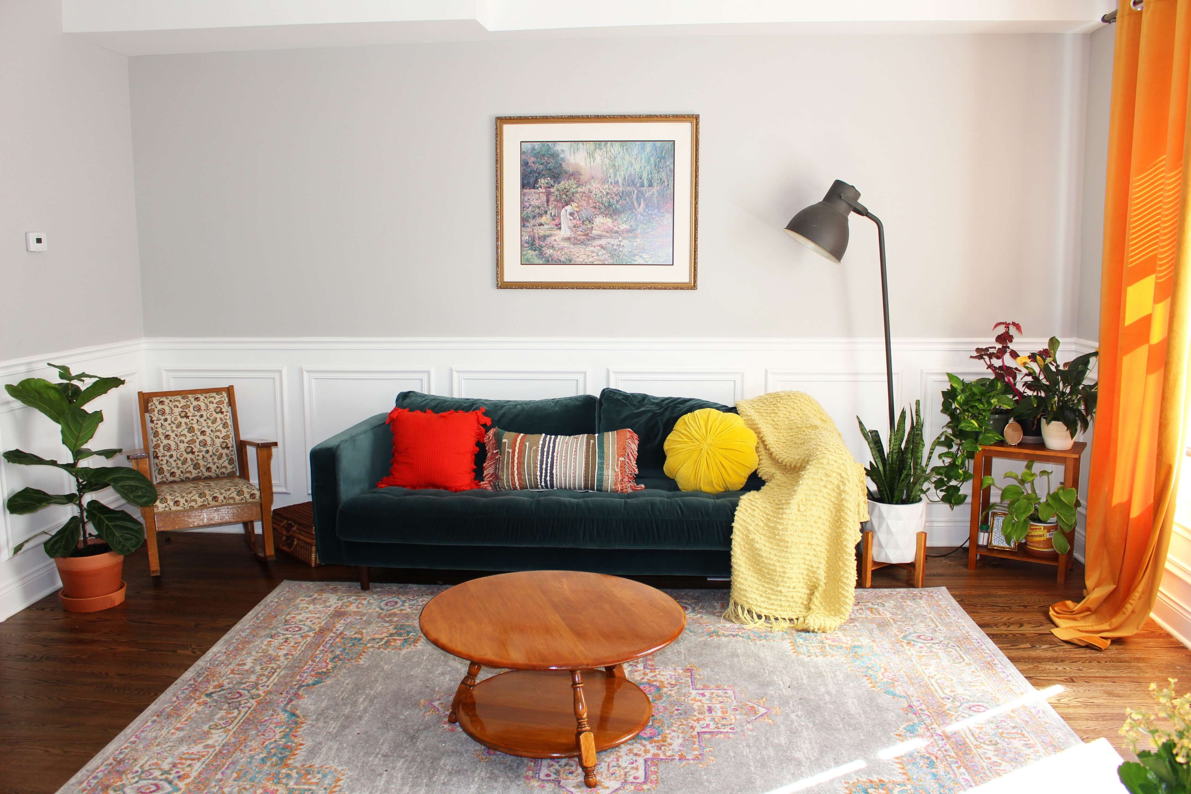 A living room features a green velvet sofa adorned with colorful pillows, a round wooden coffee table, a patterned area rug, and several plants alongside a floor lamp and framed artwork on the wall.