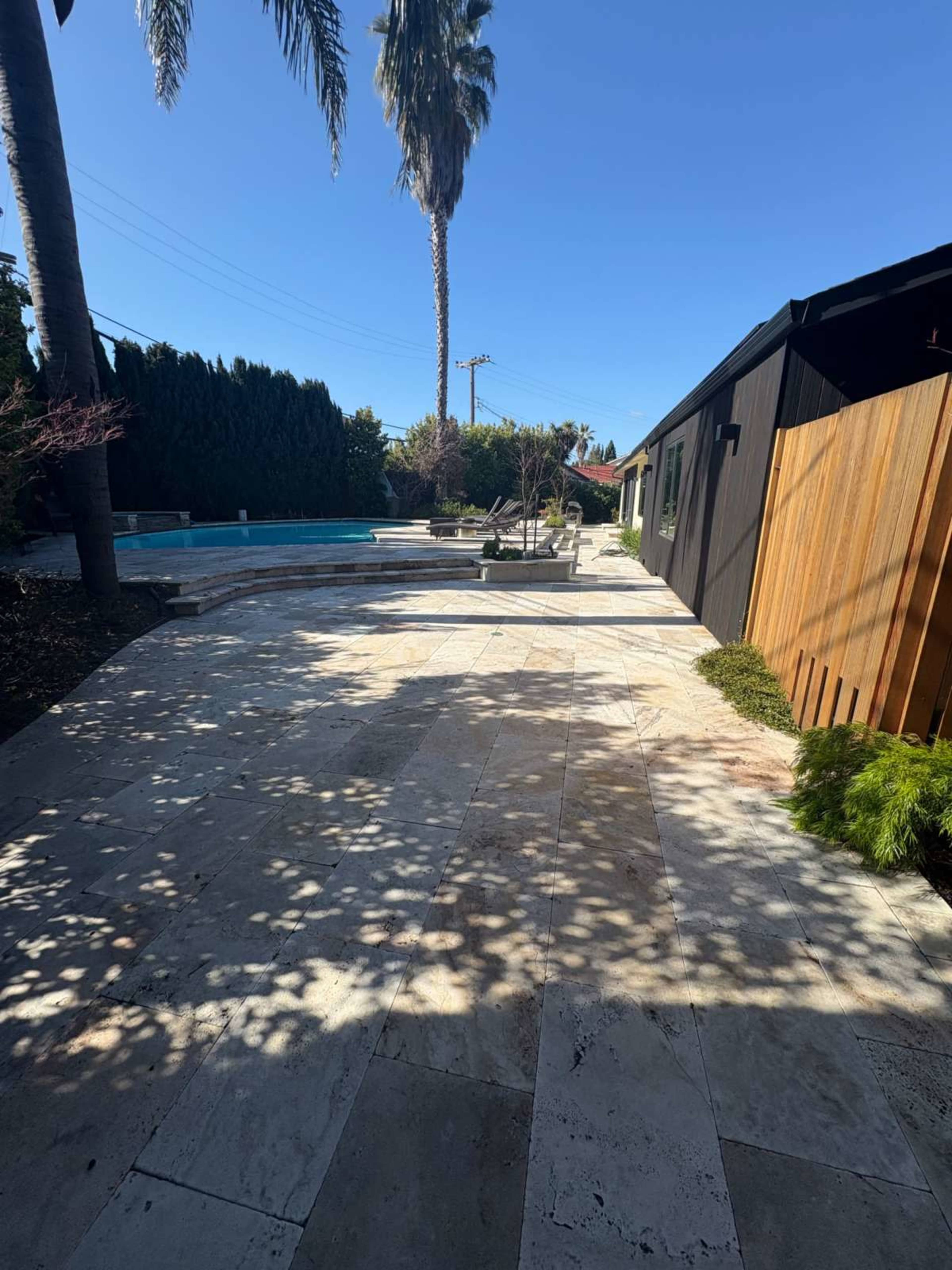 The image shows a stone-paved outdoor area next to a swimming pool, with palm trees and a modern building in the background.