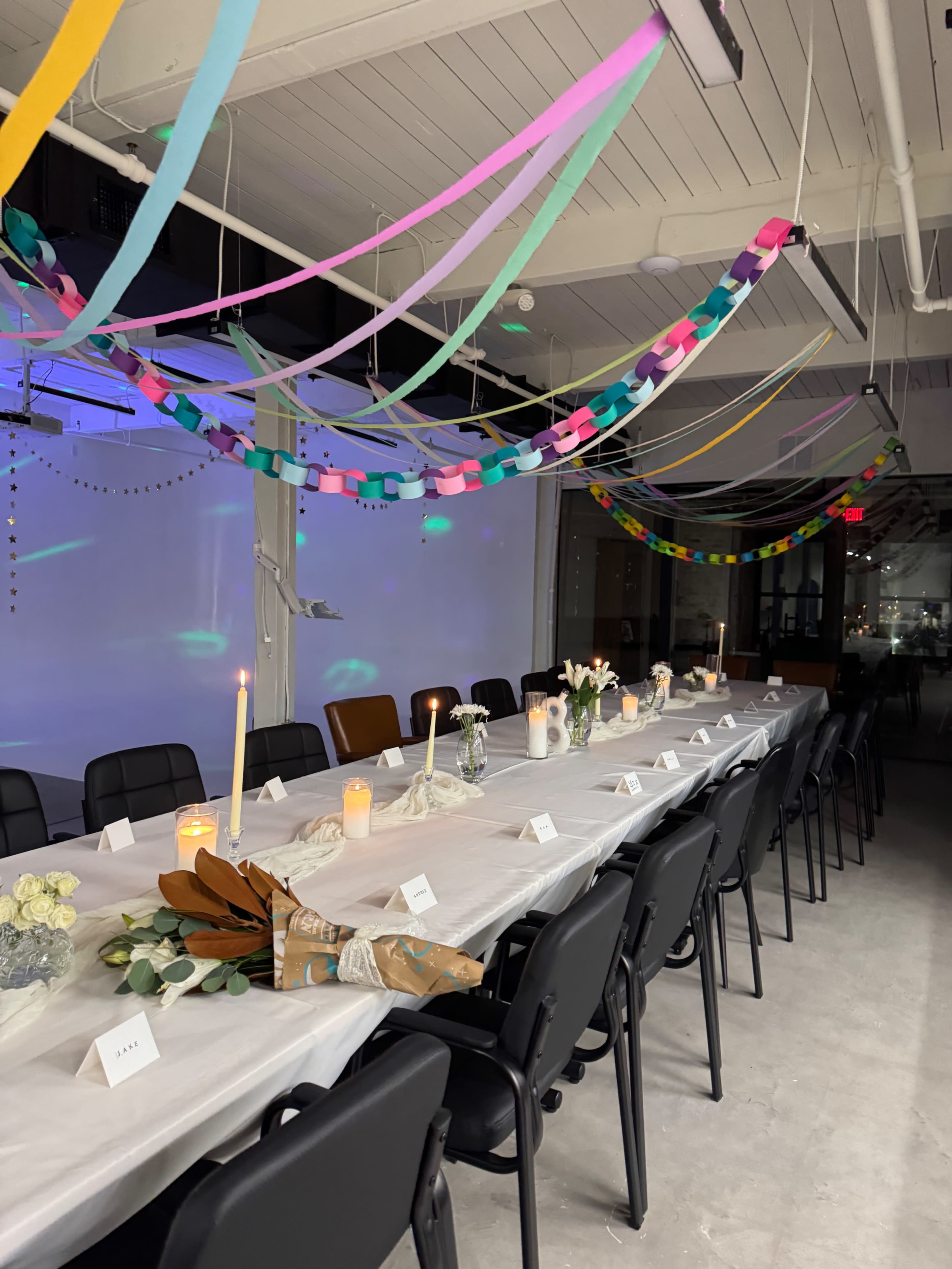 A long table is set for a gathering, decorated with candles, flowers, and colorful streamers hanging from the ceiling.