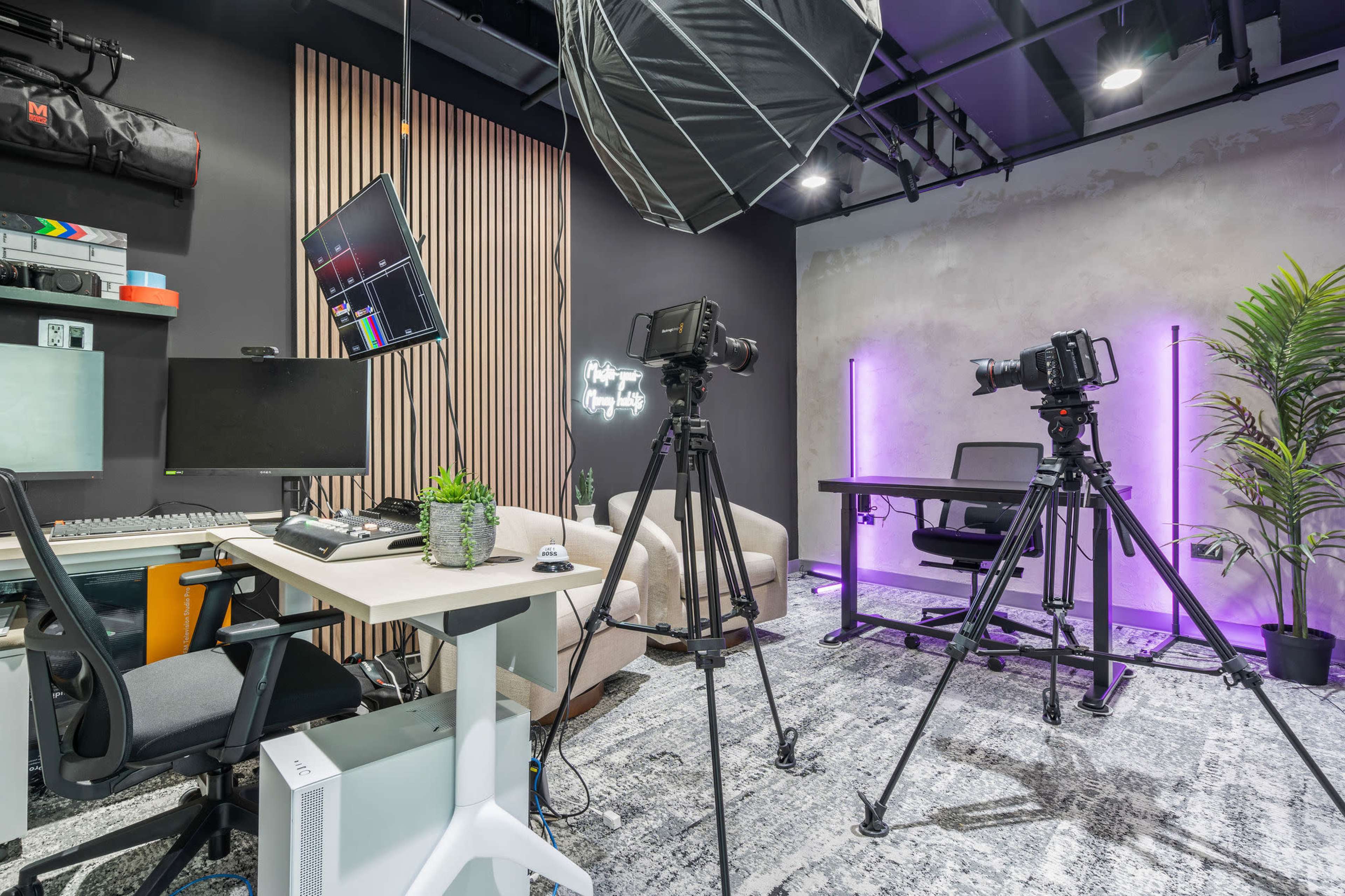 The image shows a modern studio setup with a desk, computer, cameras on tripods, and softbox lighting against a backdrop of wooden panels and a wall with neon lighting.