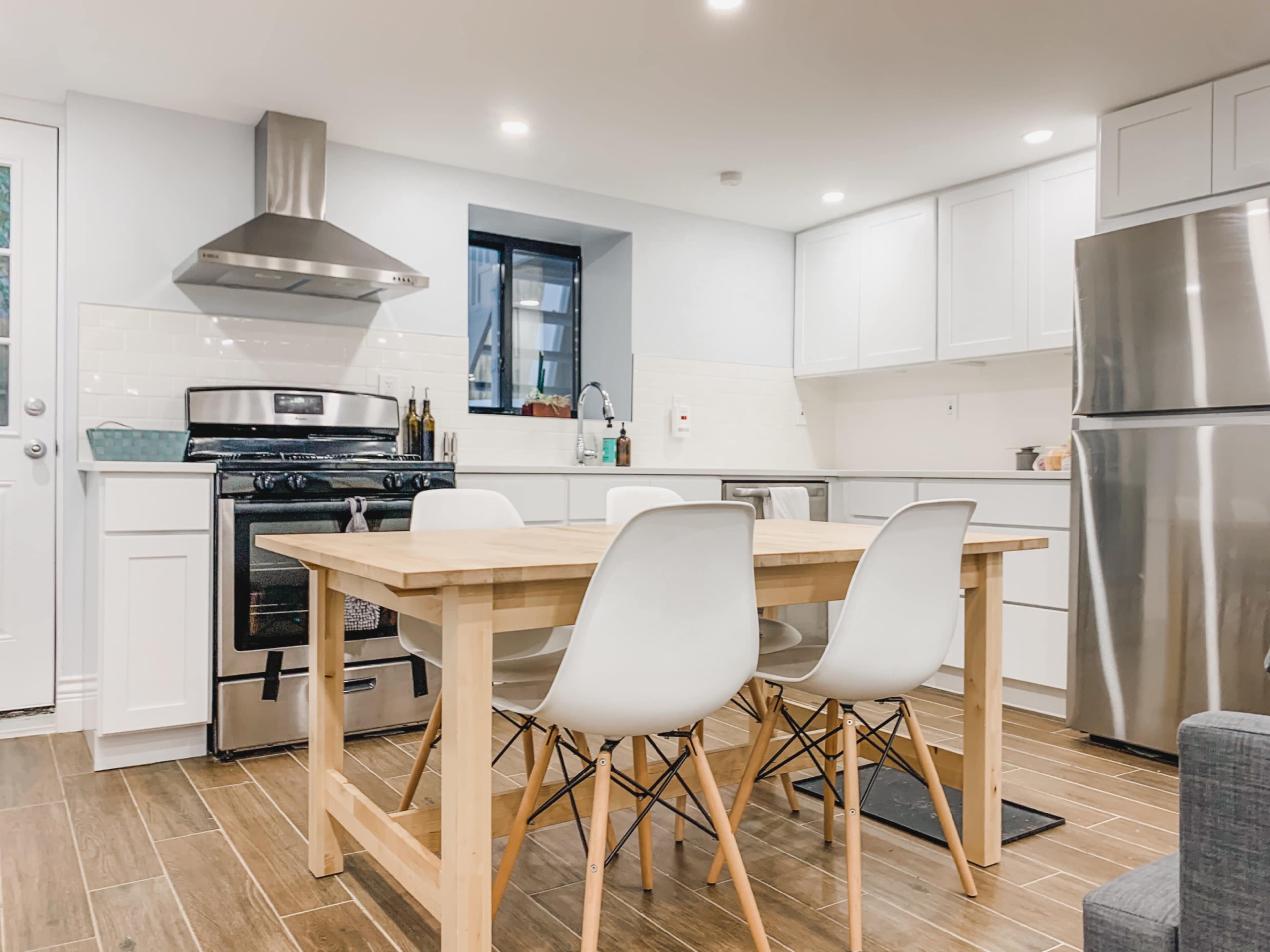 A modern kitchen features a stainless steel stove, white cabinetry, a wooden dining table with four white chairs, and a sleek refrigerator.