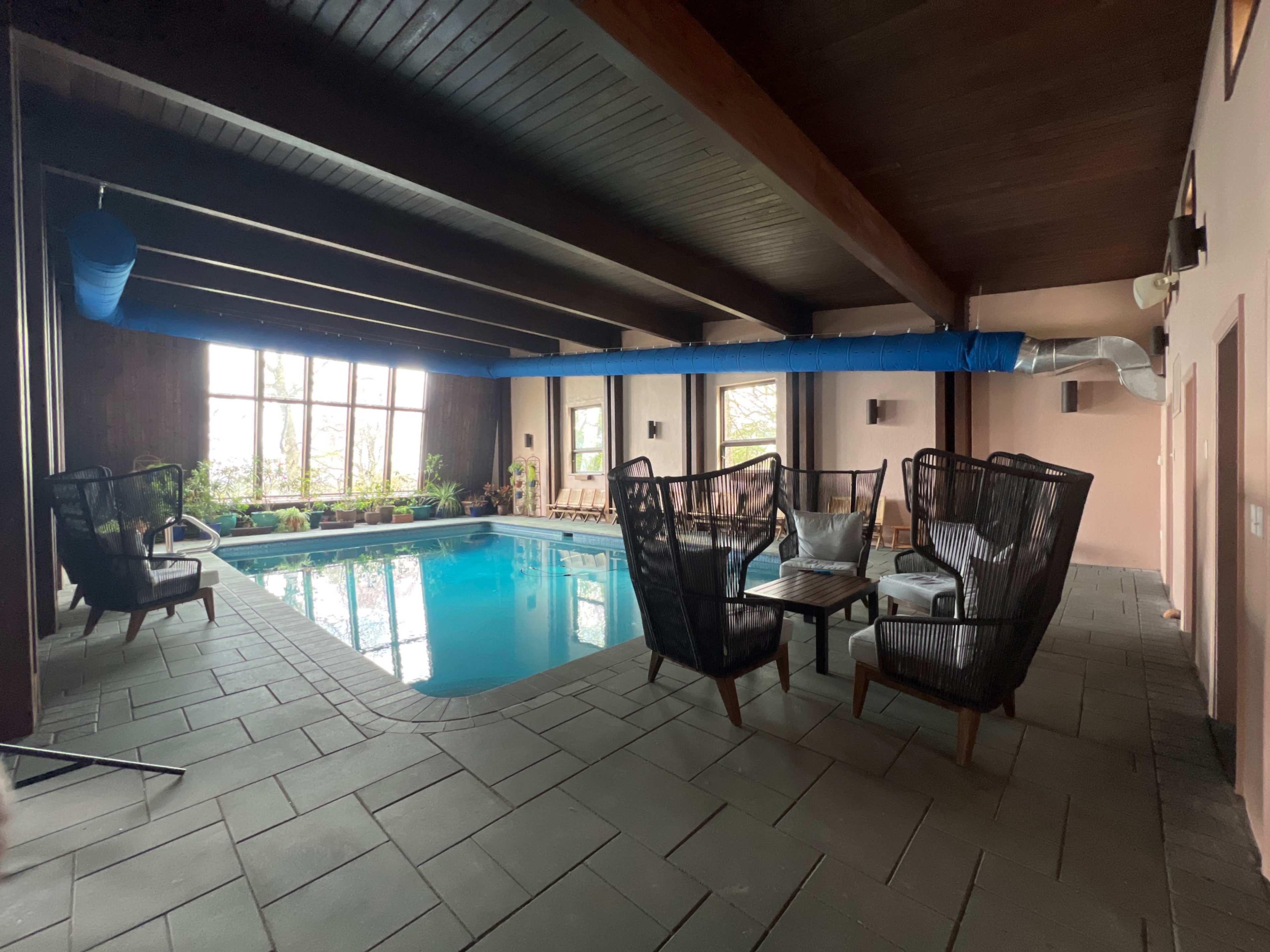 The image shows an indoor swimming pool surrounded by seating areas and large windows that allow natural light to enter.