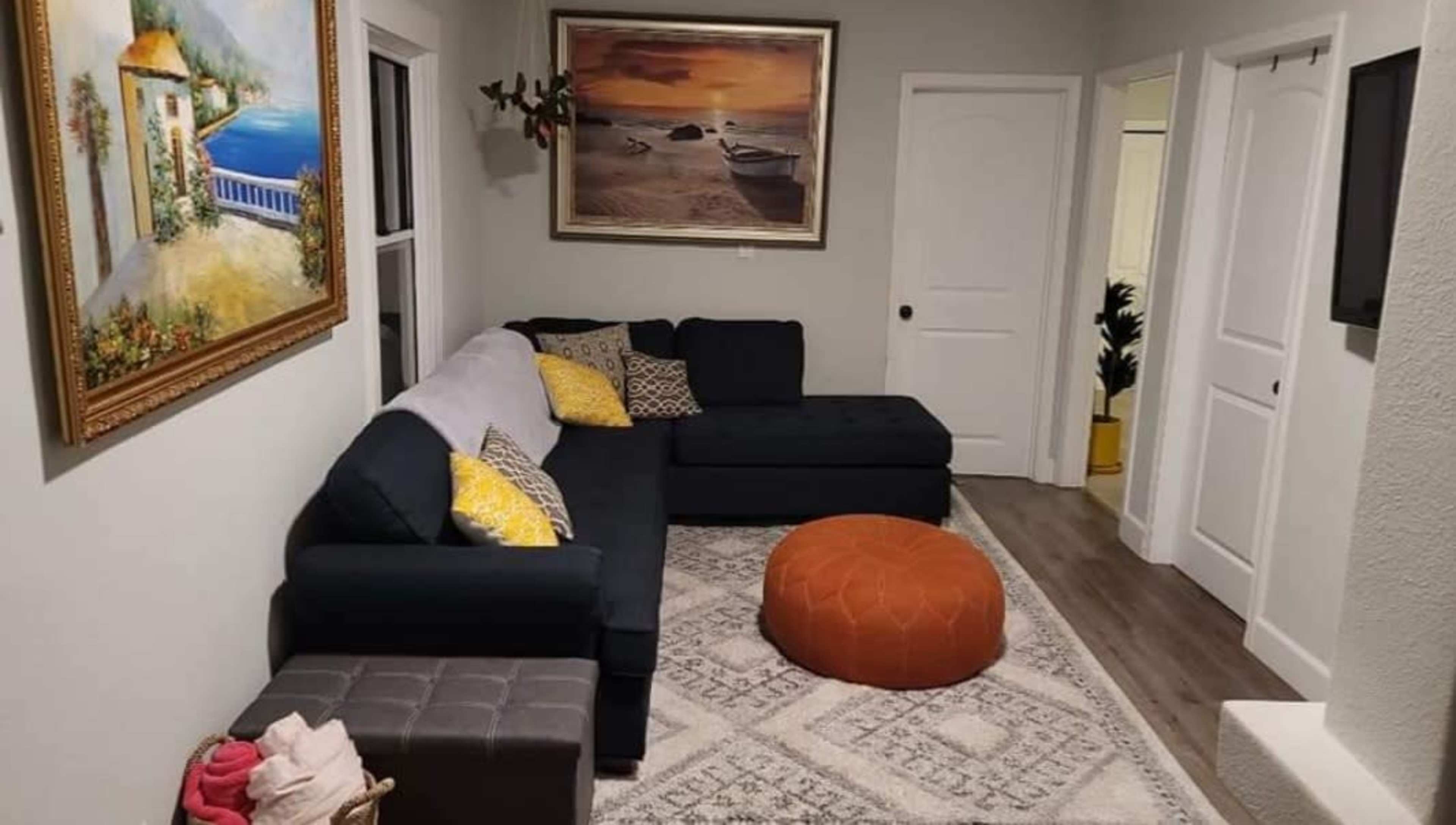 The image shows a living room with a dark sectional couch, an orange pouf, a decorative painting on the wall, and a light-colored area rug.