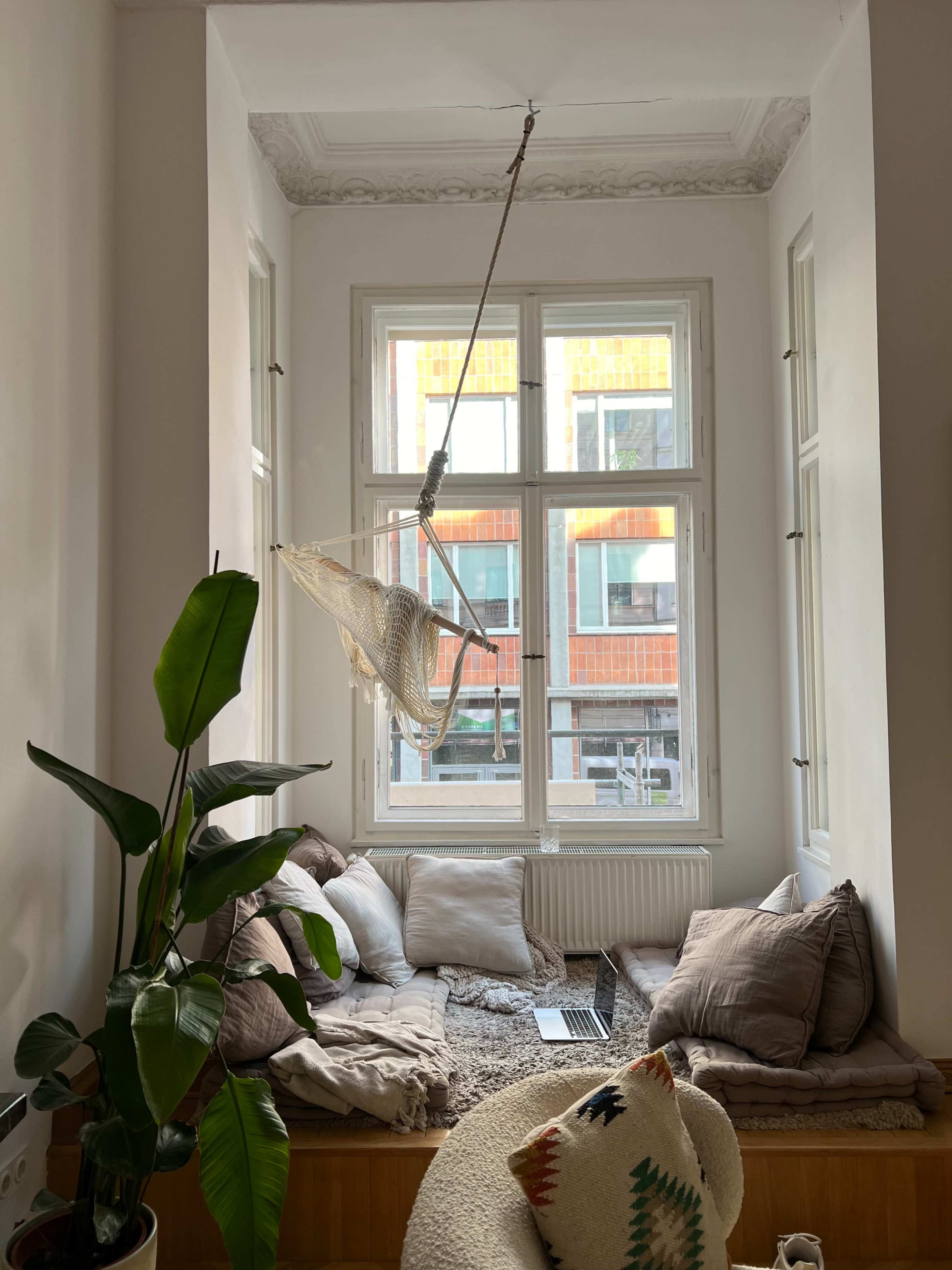 A cozy living space features a hammock chair suspended near a large window, surrounded by cushions and a potted plant.