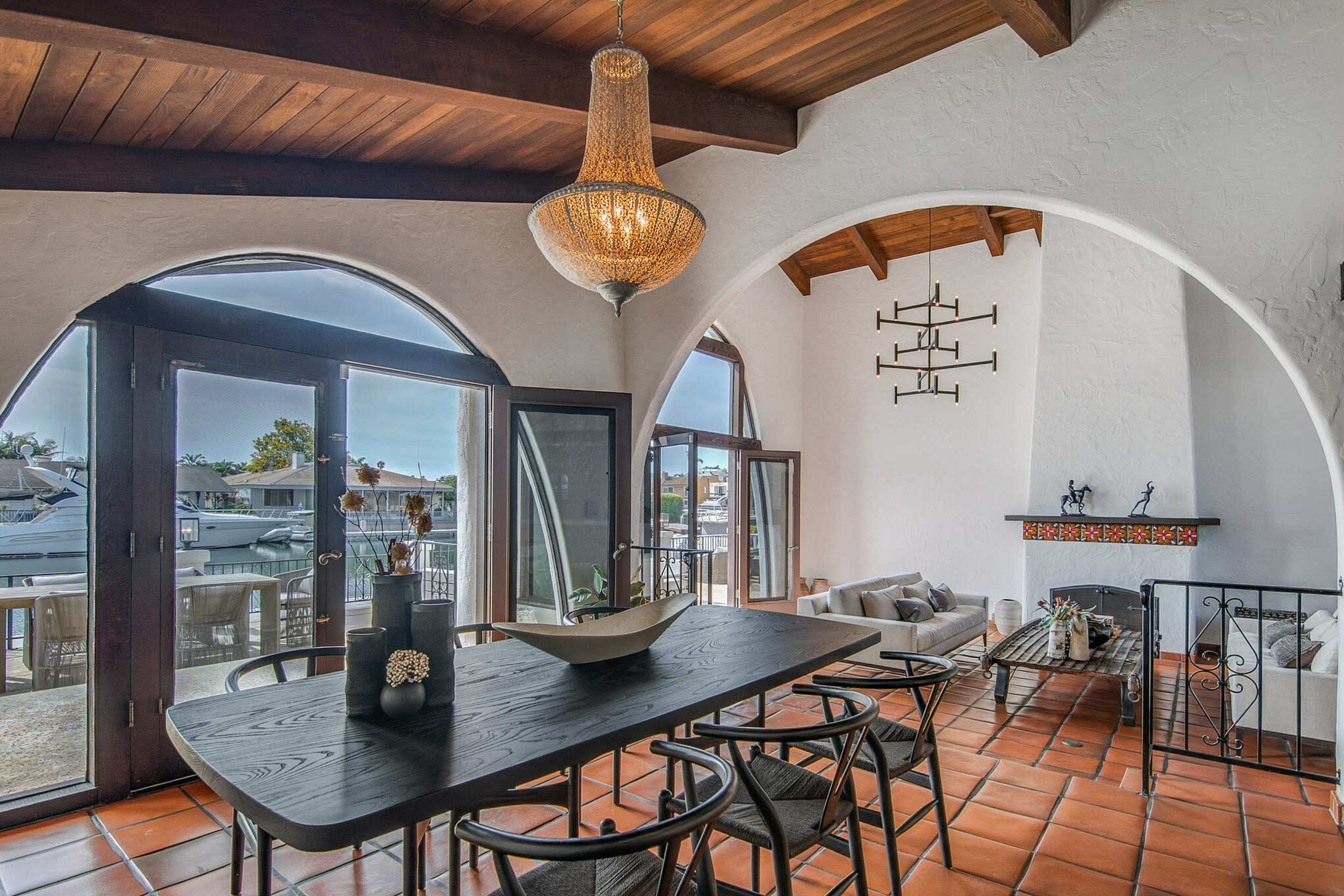 The image shows a dining area with a large black table and curved archways, featuring a chandelier and views of an outdoor space through large glass doors.