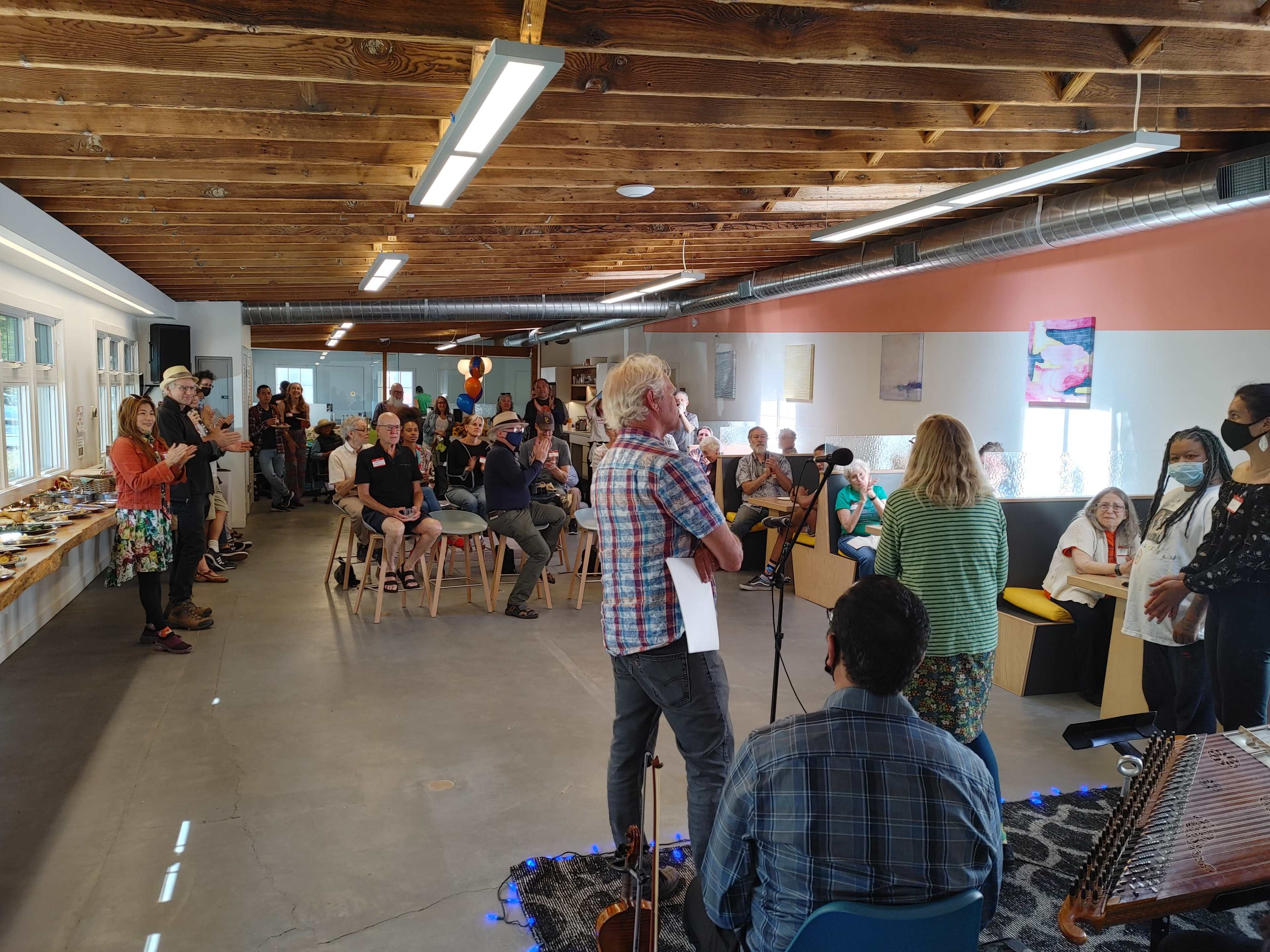 A crowd of people is seated and standing in a spacious community room while a performer sings and plays guitar on a small stage.