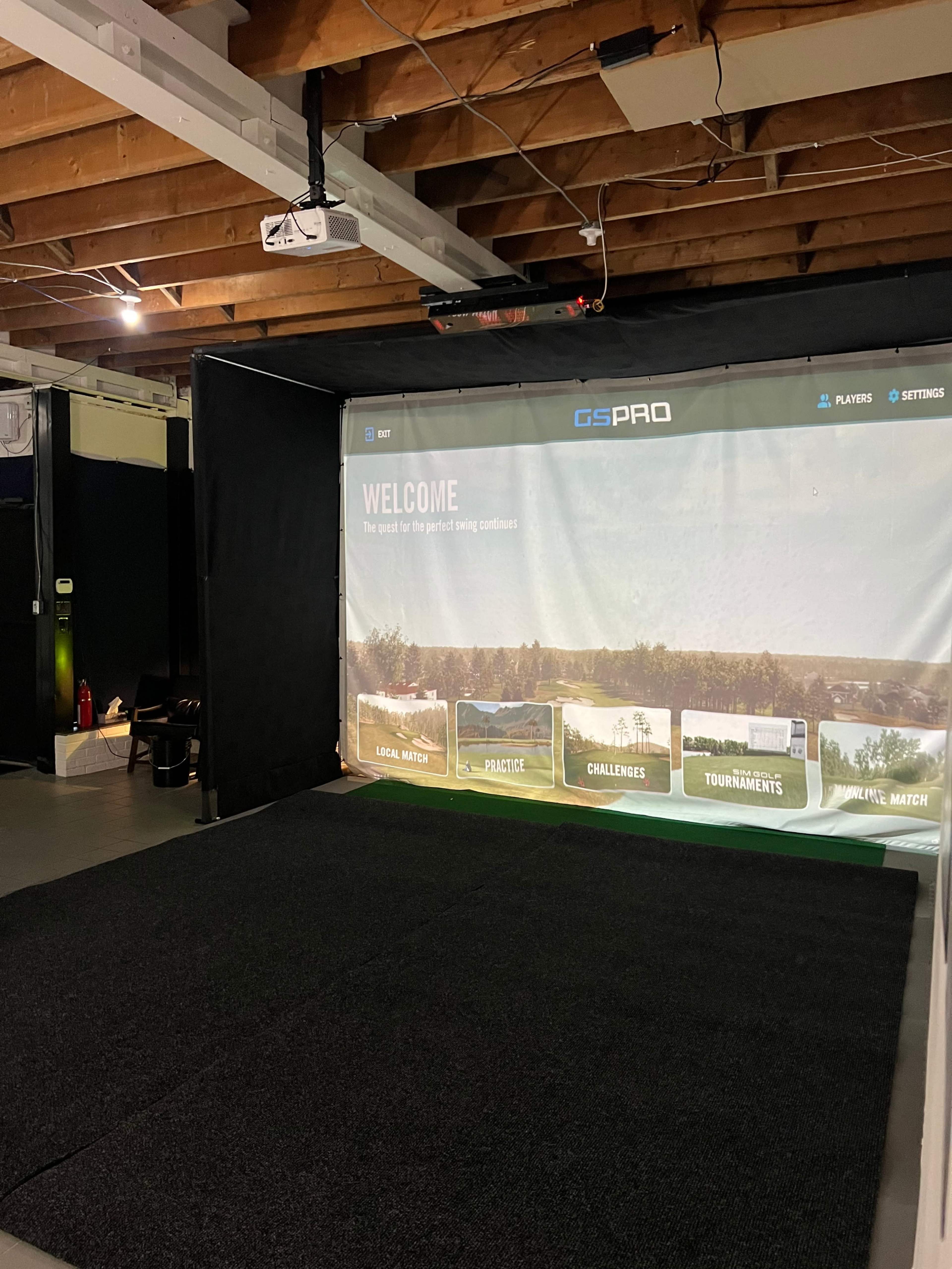 The image shows a golf simulator setup in a basement, featuring a large screen displaying a welcome message and various options for gameplay.
