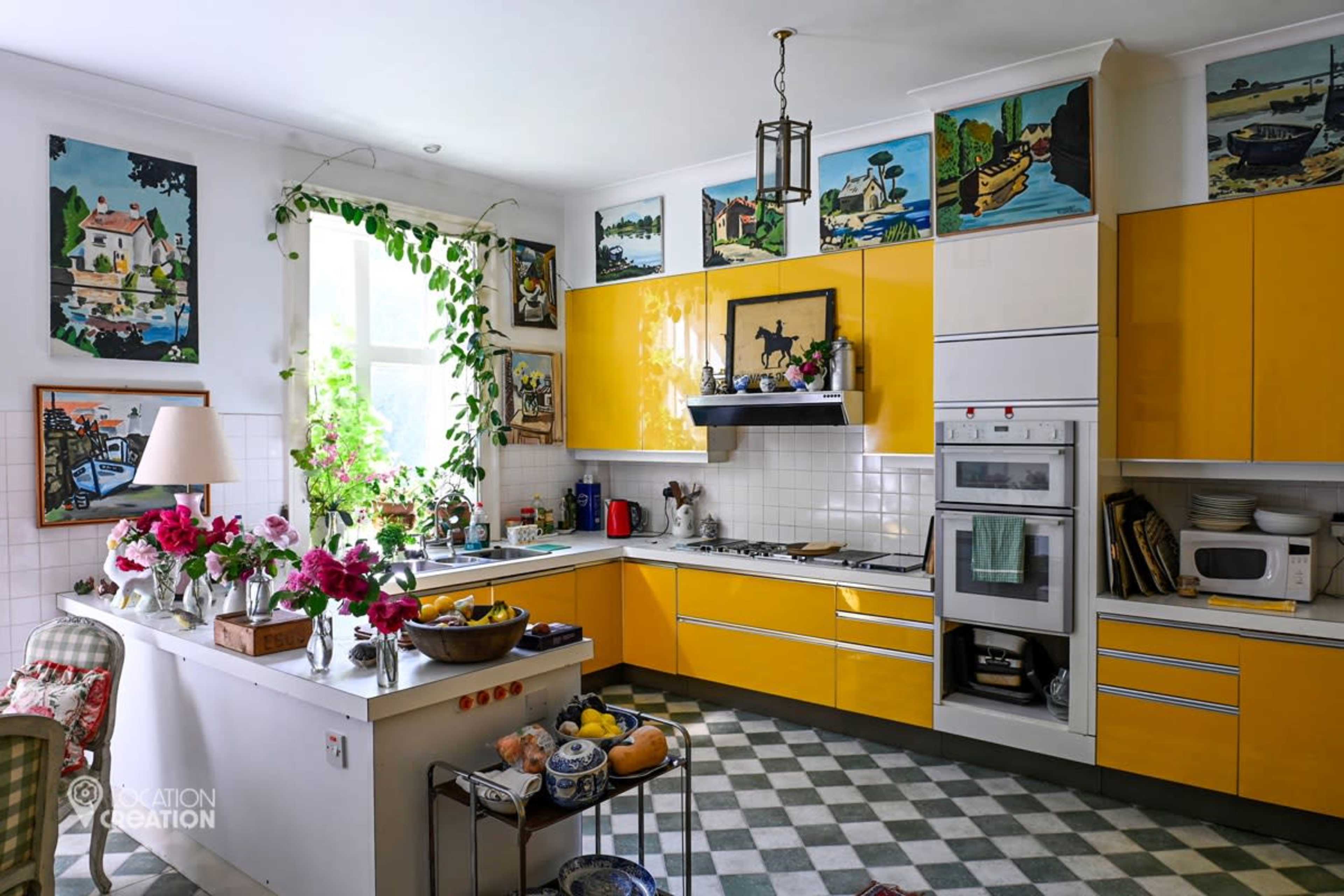 The image shows a bright kitchen with yellow cabinets, a checkered floor, and various plants and flowers on the counter.