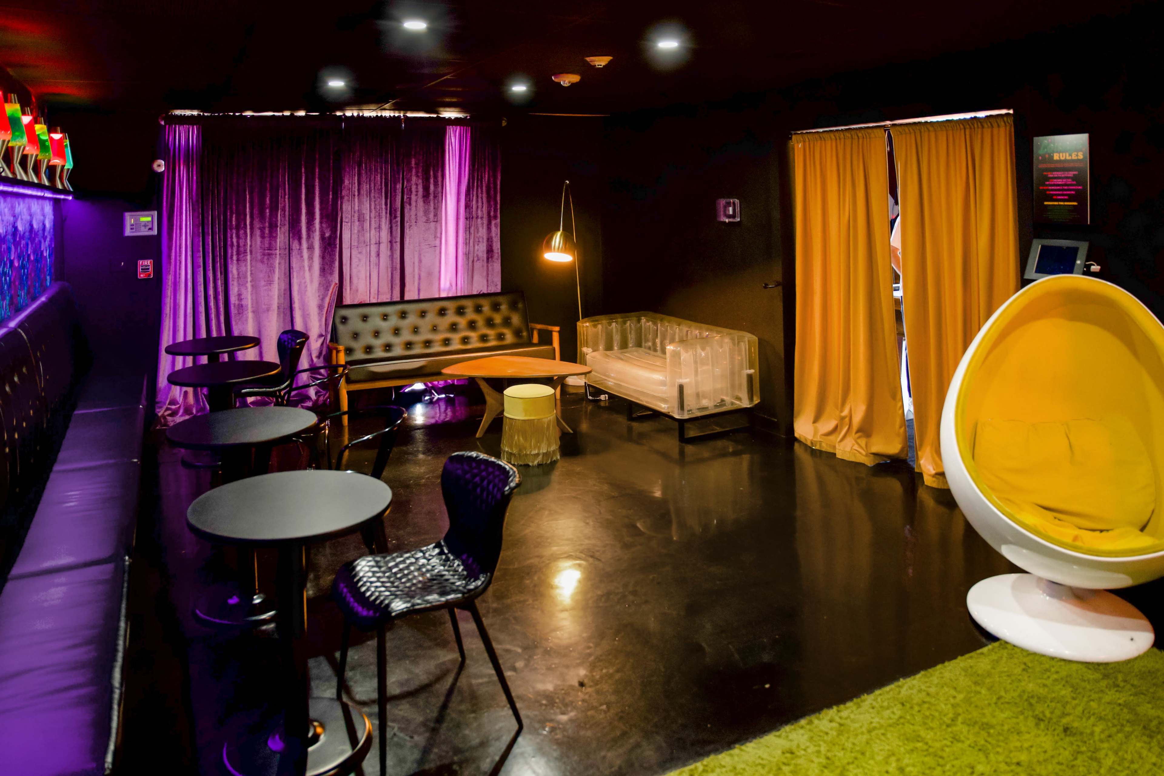 The scene features a dark lounge with a mix of seating options, including a yellow egg chair, sleek black tables, and a glass coffee table, illuminated by colorful lighting.
