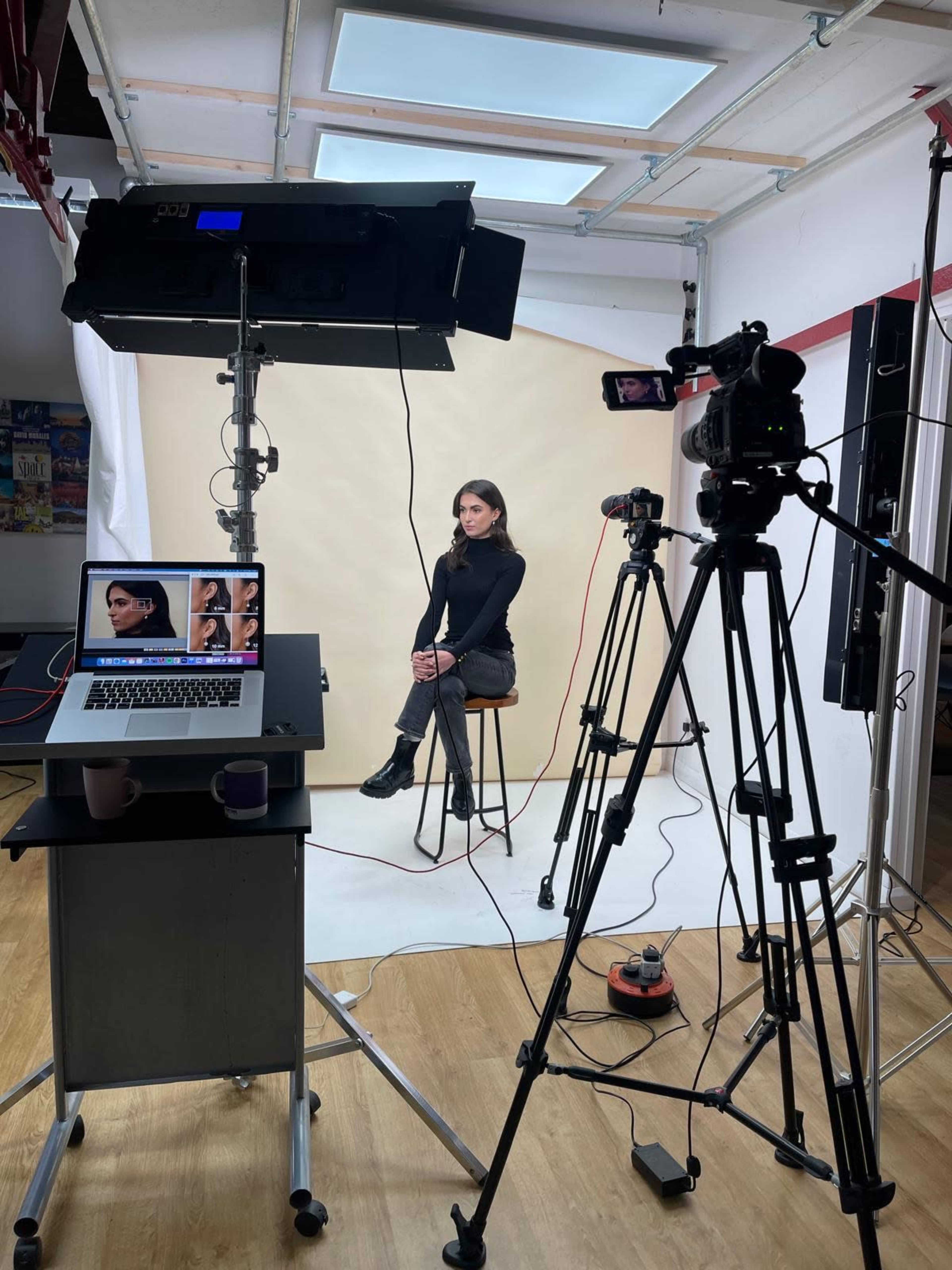 A person sits on a stool in front of a beige backdrop, surrounded by video and lighting equipment in a photography studio.