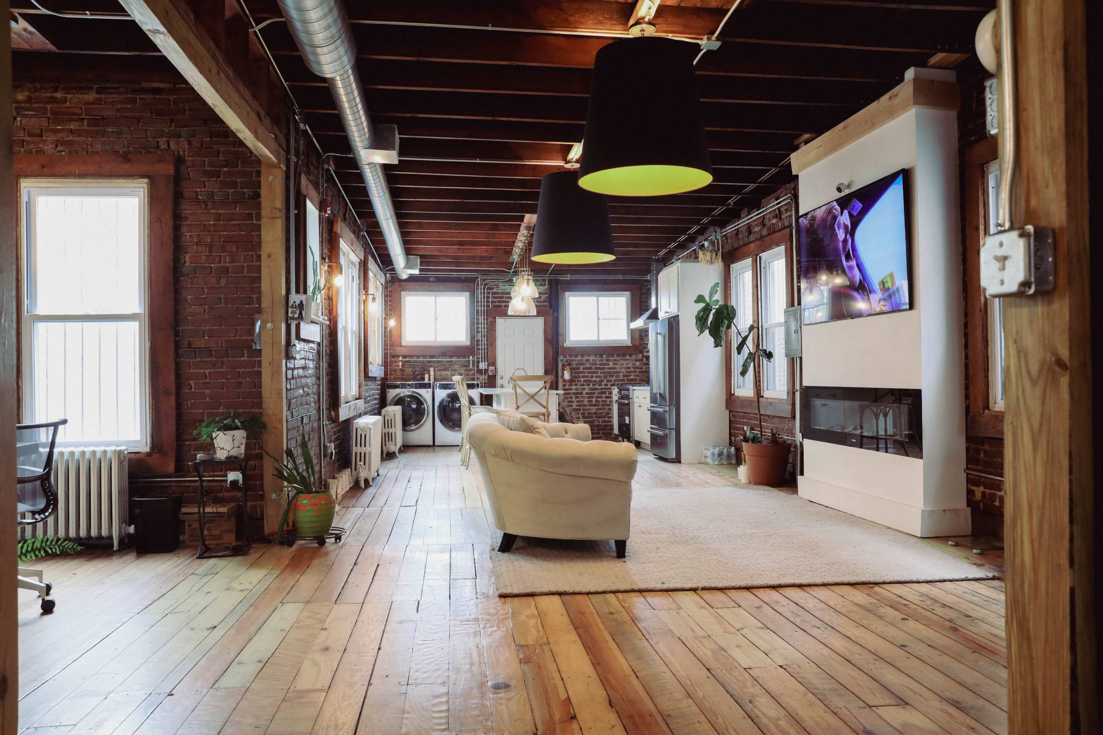 A spacious loft-style living area featuring exposed brick walls, wooden beams, and a couch facing a mounted television.