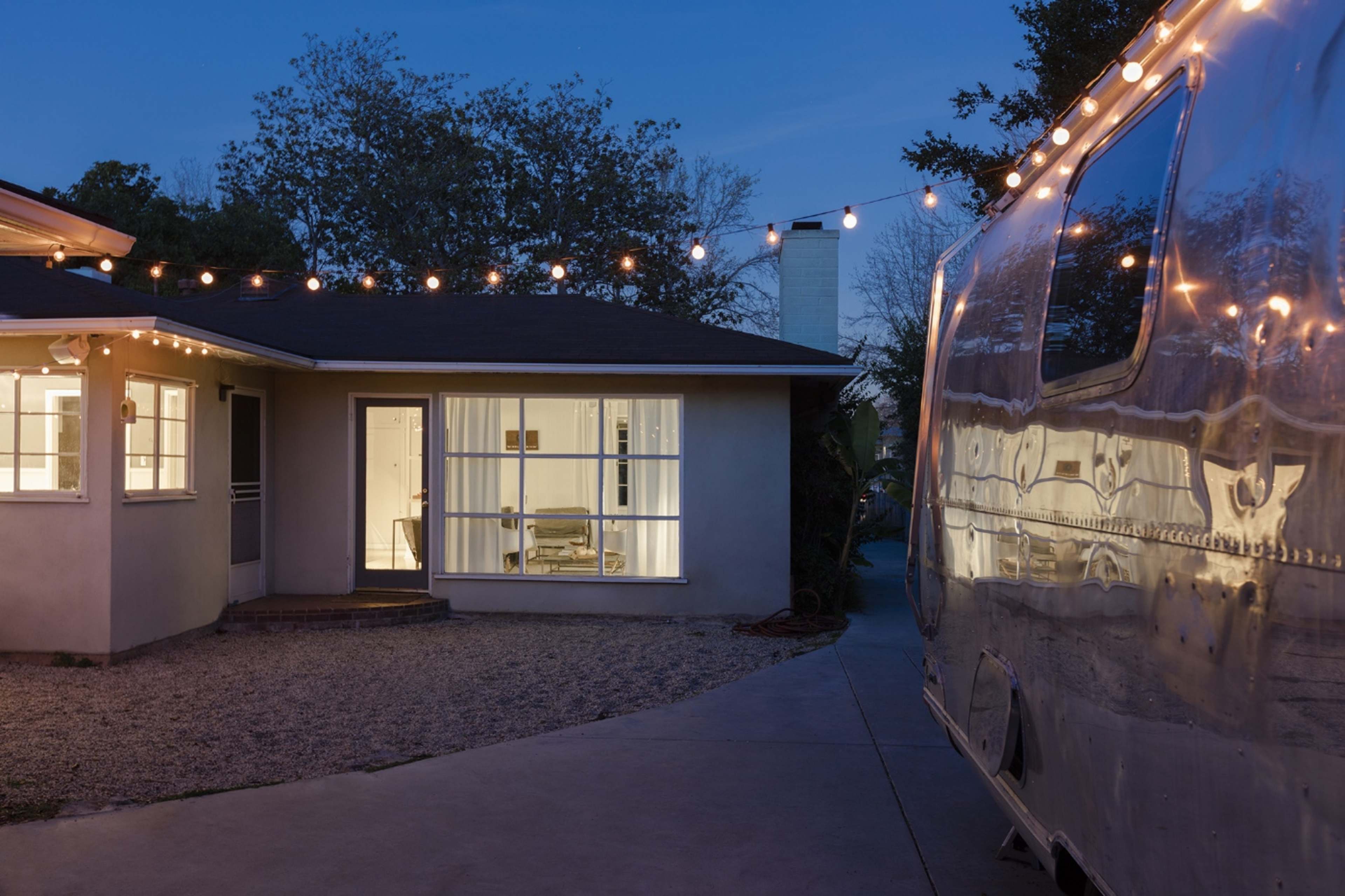 A brightly lit house with large windows stands beside a parked silver trailer, illuminated by string lights in the evening sky.