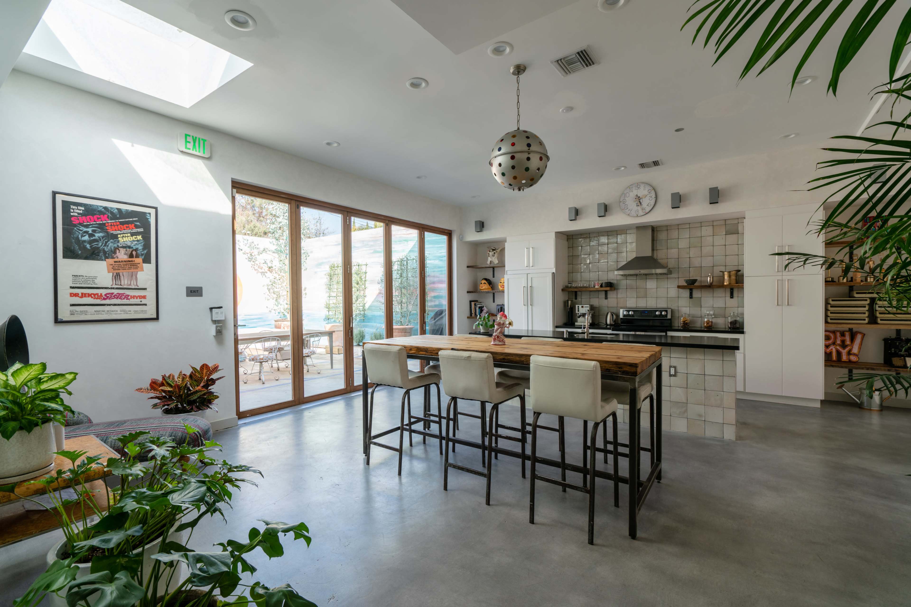 A modern kitchen and dining area featuring a large wooden table with bar stools, a gas stove, and large glass doors opening to an outdoor space, surrounded by potted plants.