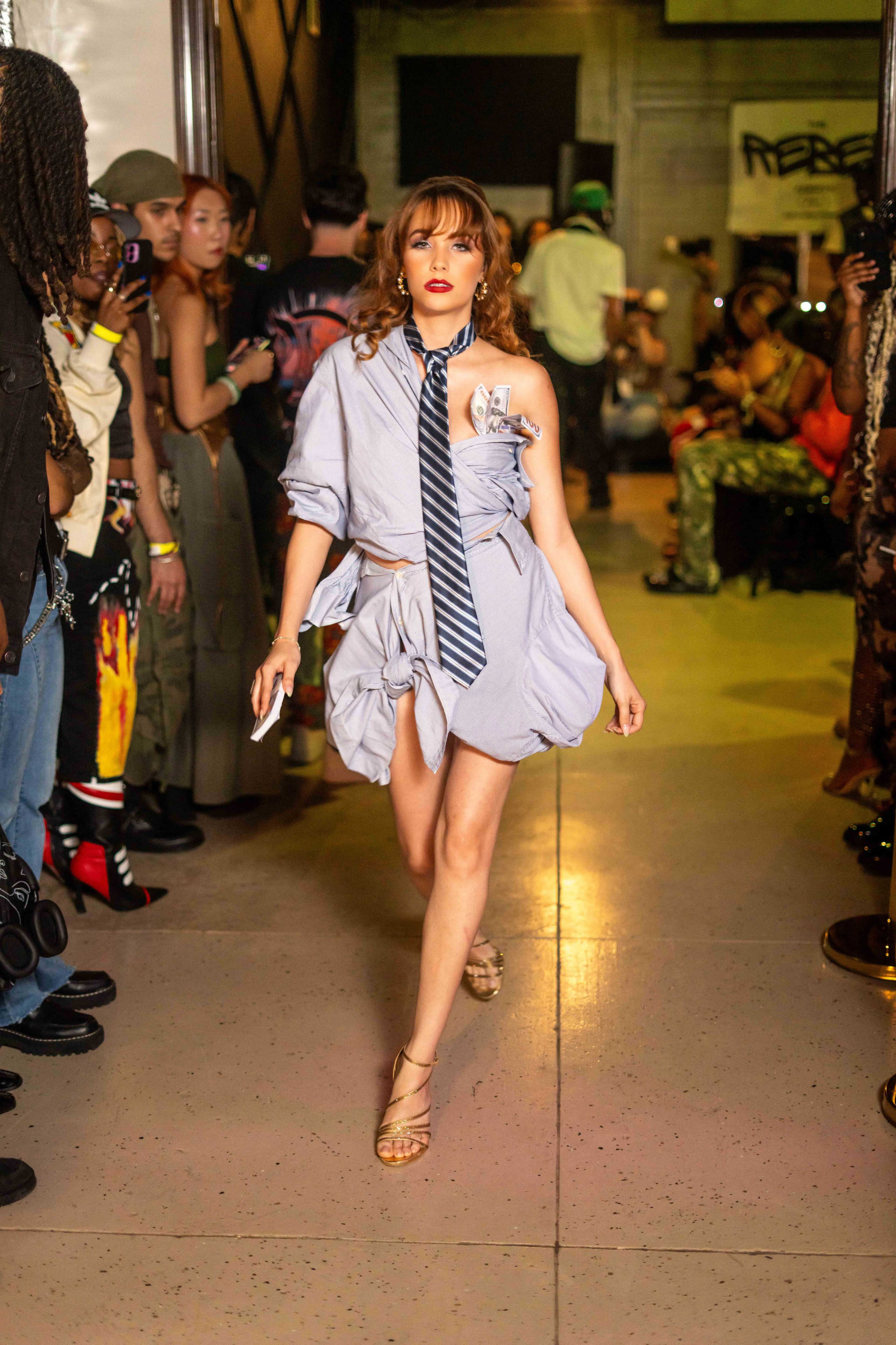 A model walks down a runway wearing a partially buttoned shirt and a tie, surrounded by an audience at a fashion event.