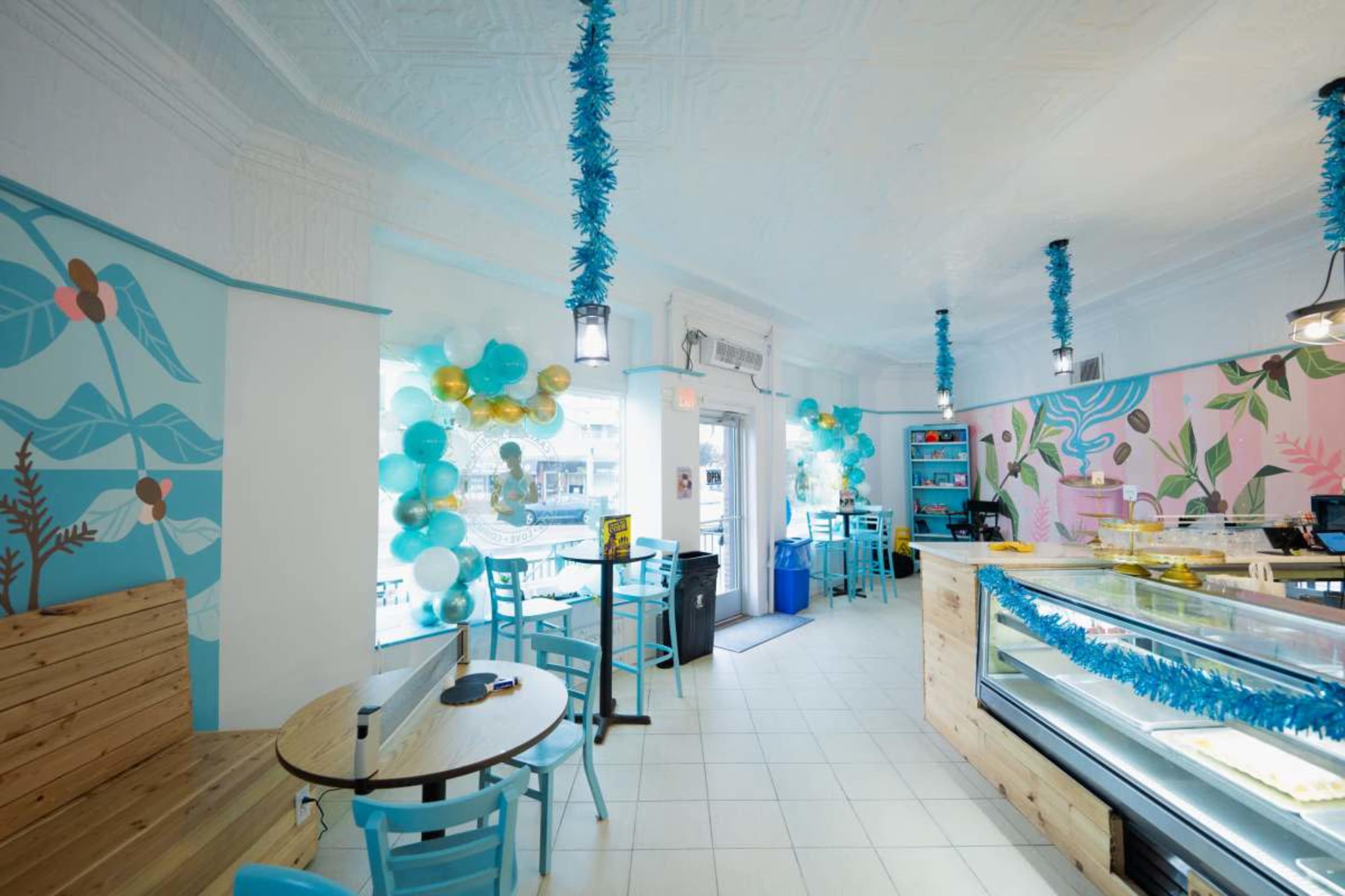 The interior of a brightly decorated café features blue and gold balloons, colorful wall murals, and light wooden furniture.