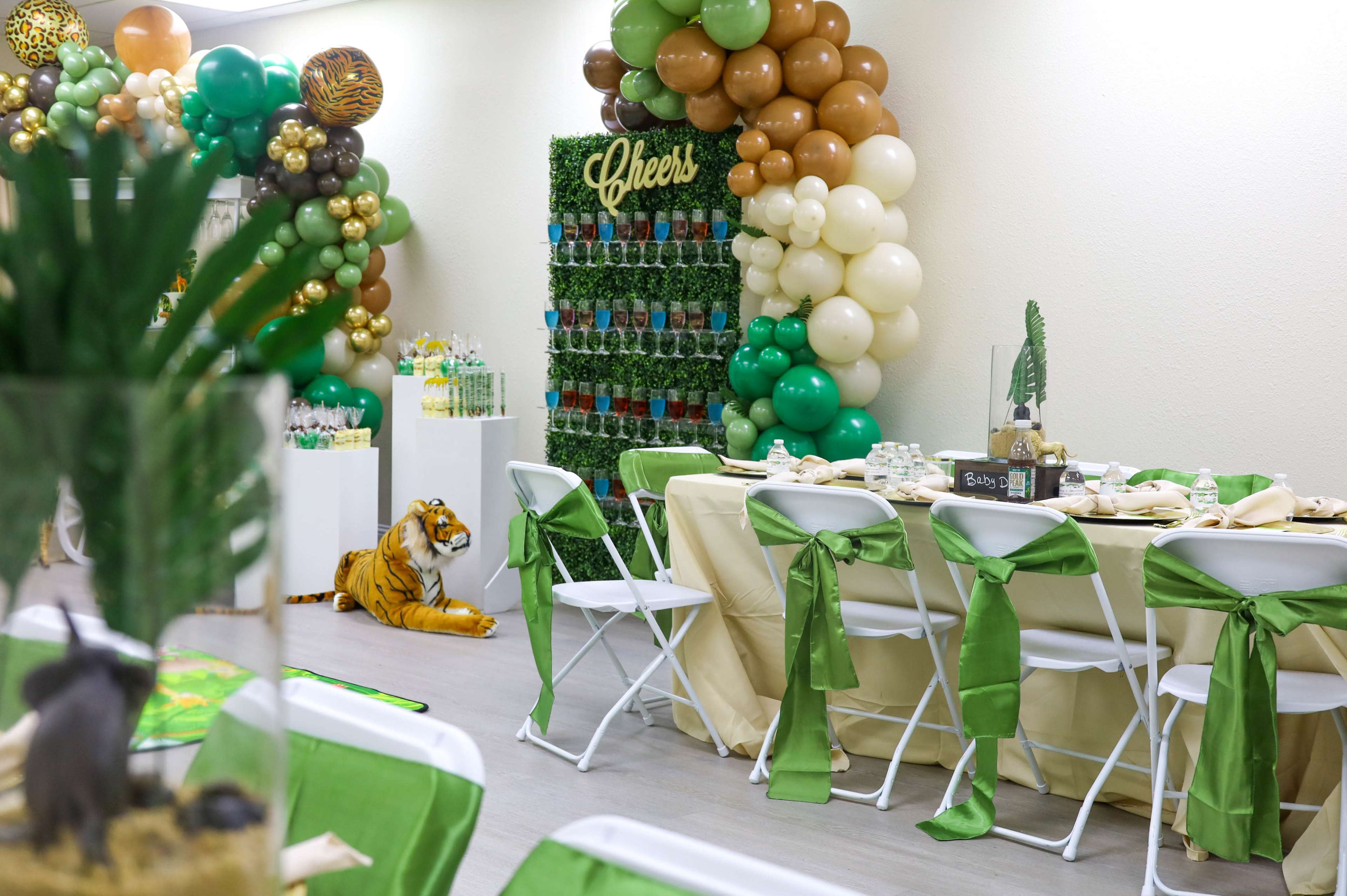 A party setting with a decorated table, green tablecloths, a wall of balloons, and a large display of drinks, along with a tiger decoration on the floor.