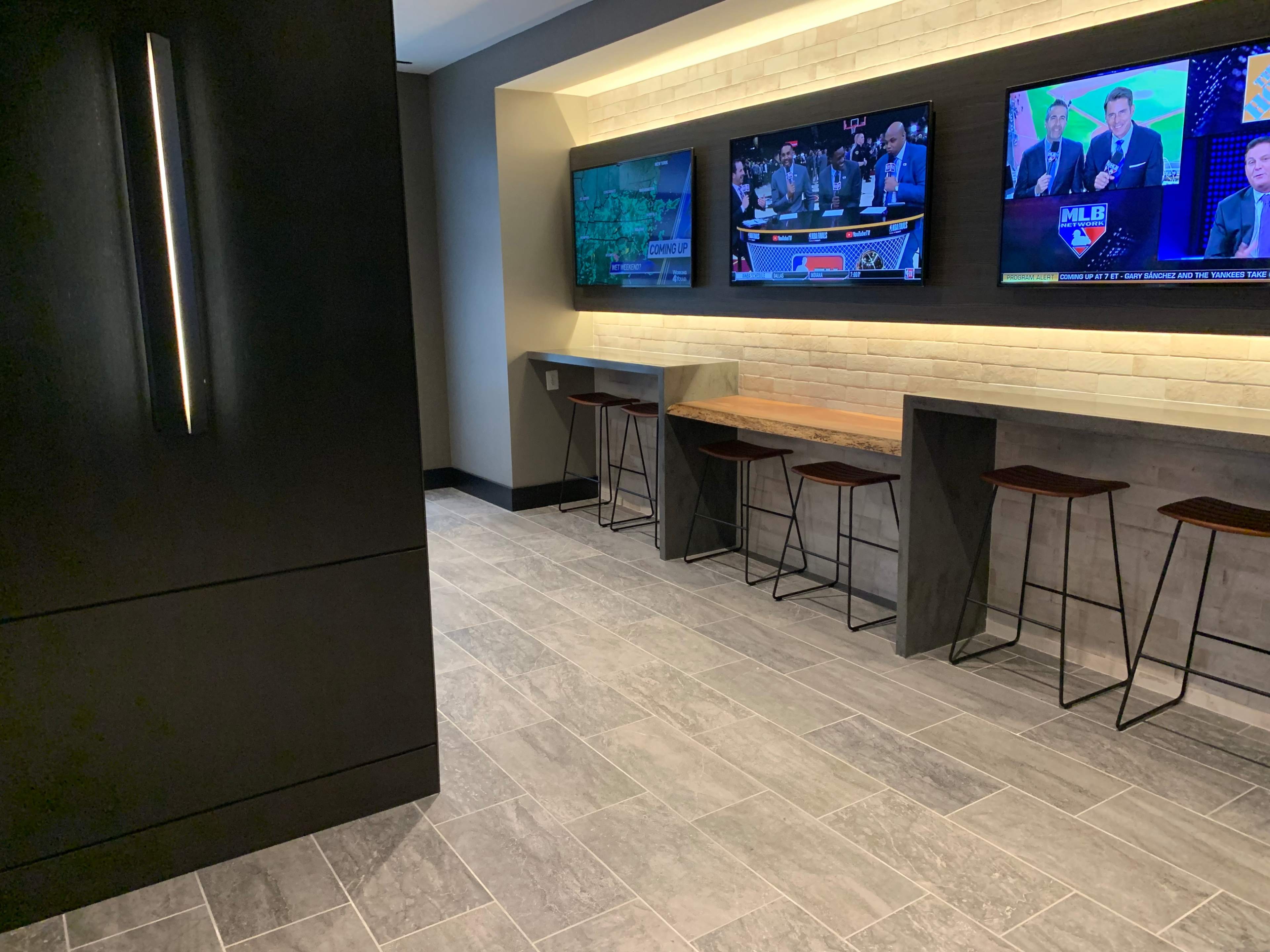 A modern lounge area featuring multiple flat-screen TVs mounted on a wall and sleek bar stools lined up along a counter.
