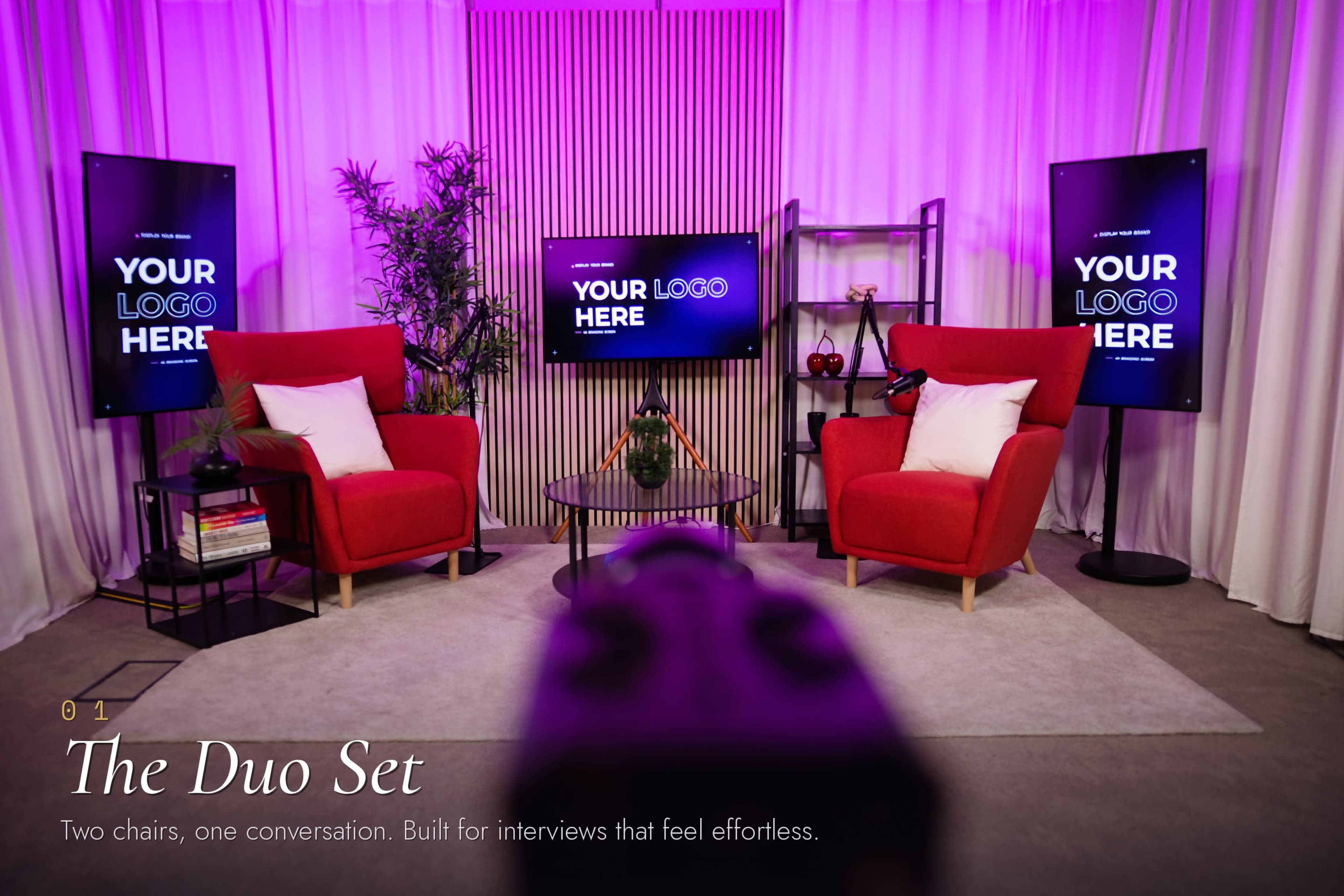 The image shows a modern interview setup with two red chairs facing each other, flanked by two screens displaying "YOUR LOGO HERE," and surrounded by decorative plants and a coffee table.