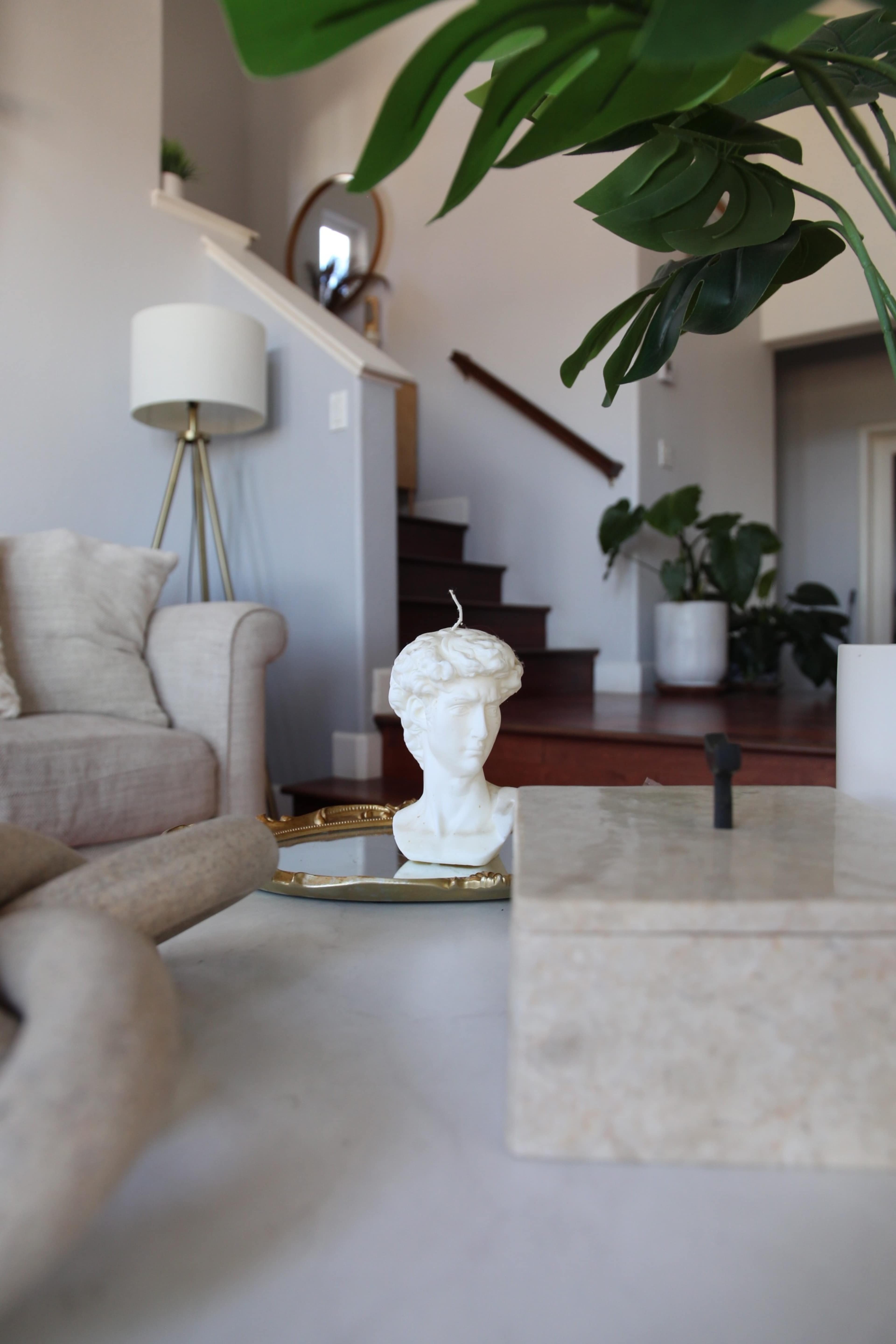 The image shows a living room with a white sculpture of a bust on a coffee table, a green plant in the foreground, and stairs leading to an upper level in the background.