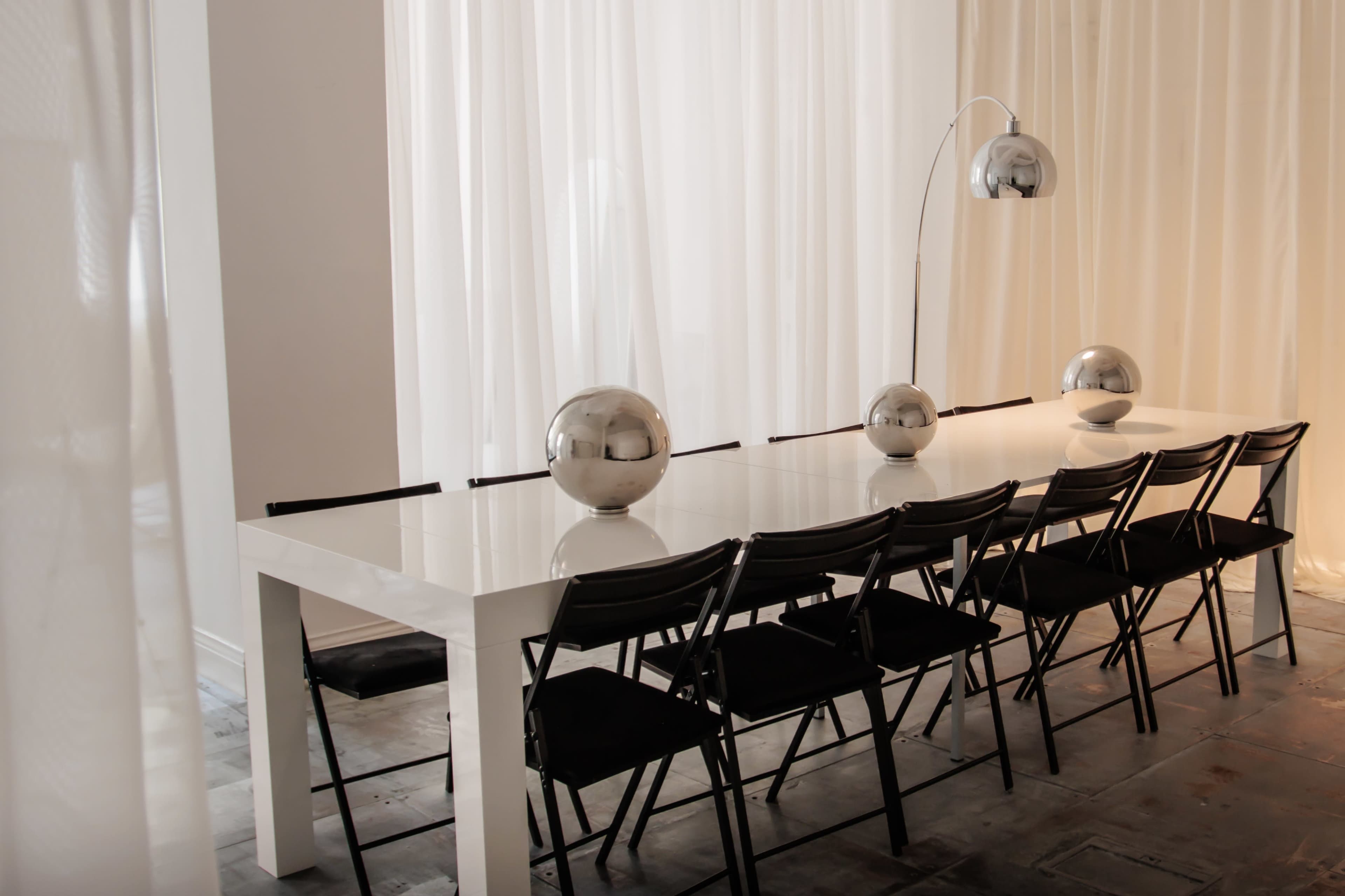 A long, white dining table is set with three metallic spheres, accompanied by black folding chairs and illuminated by an arc floor lamp.