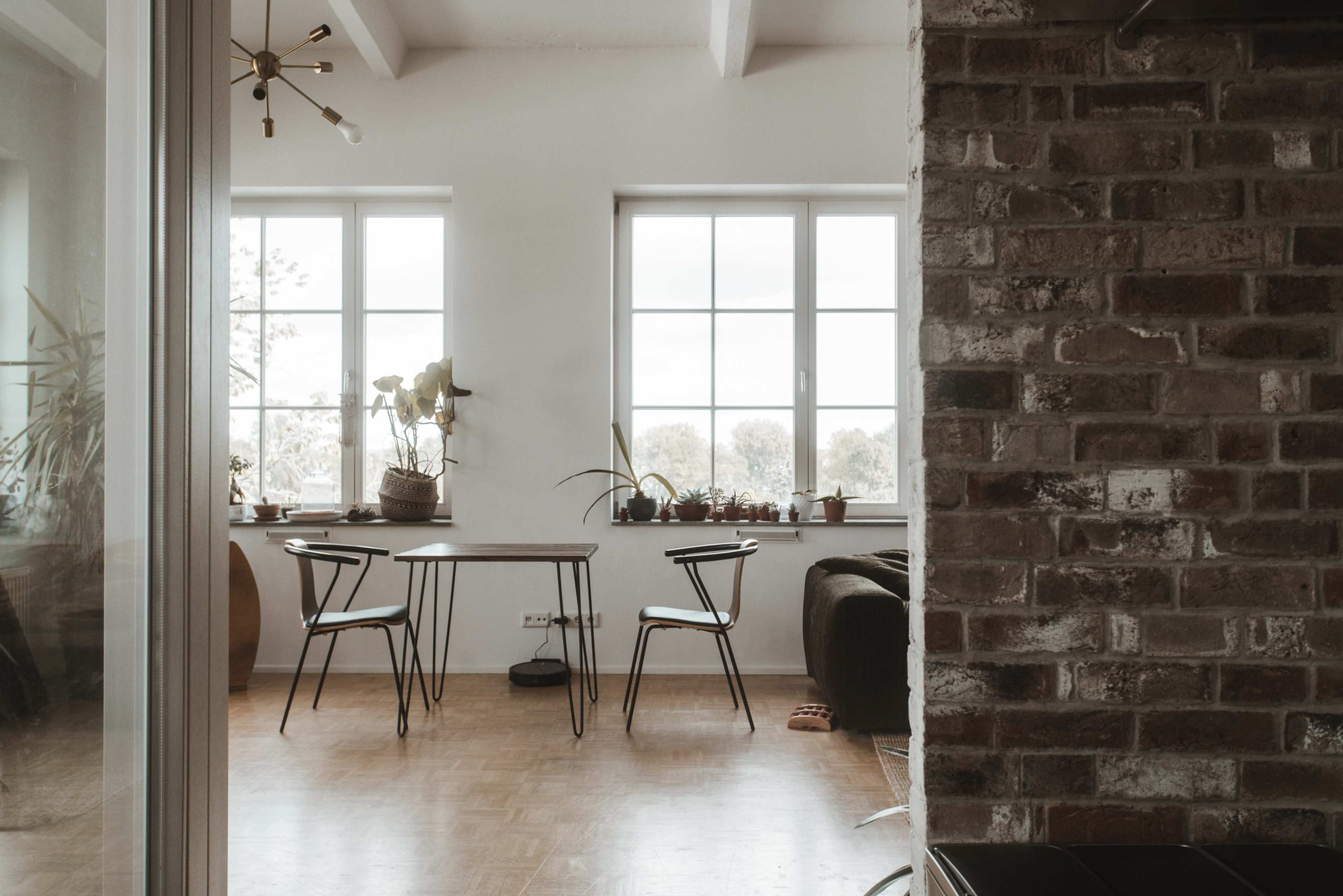 The image shows a minimalist dining area with a wooden table and two chairs facing large windows, surrounded by various potted plants.