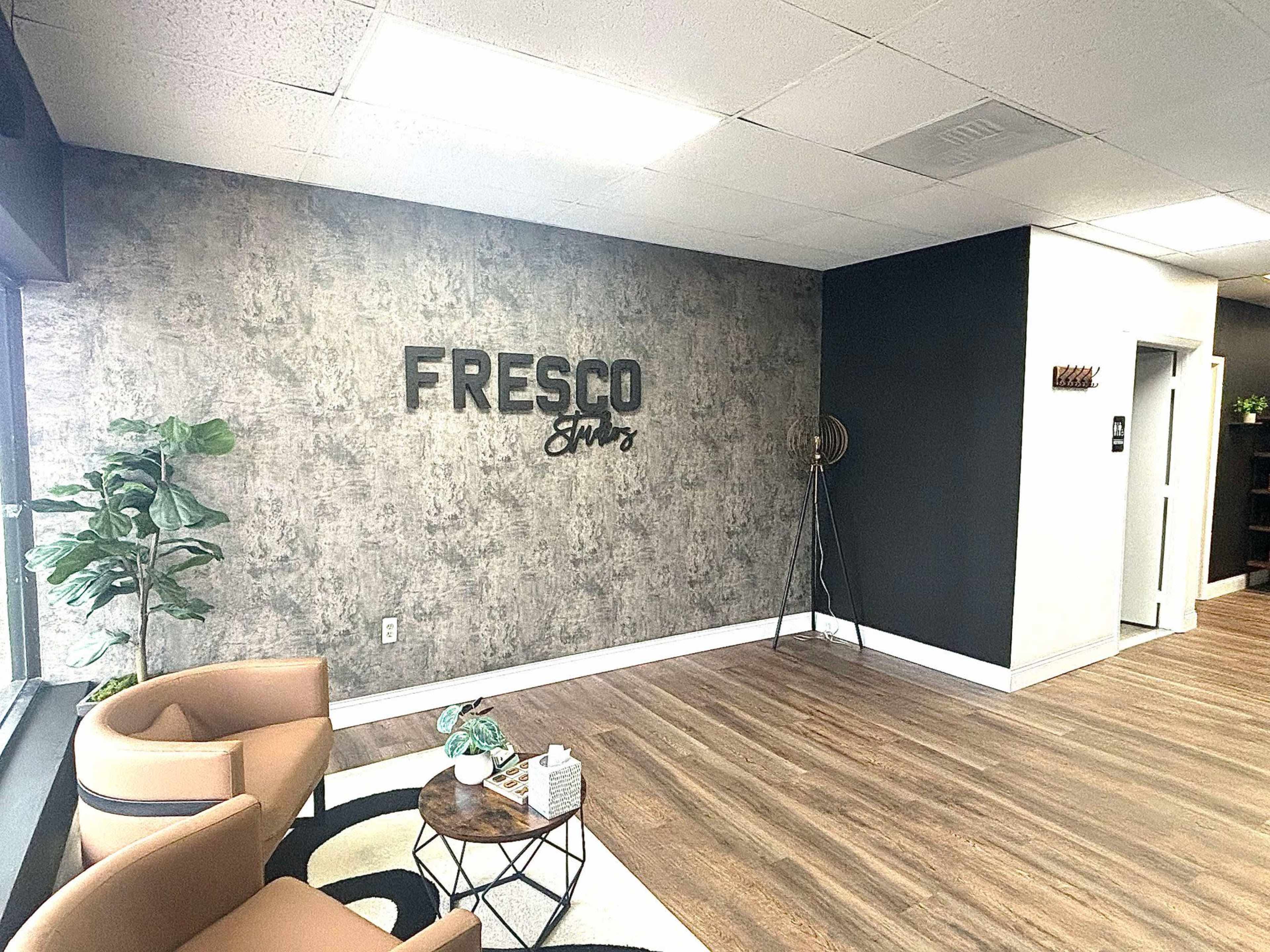 The image shows a modern reception area with a textured wall featuring the word "FRESCO" and minimalistic furniture, including two tan chairs and a small table.