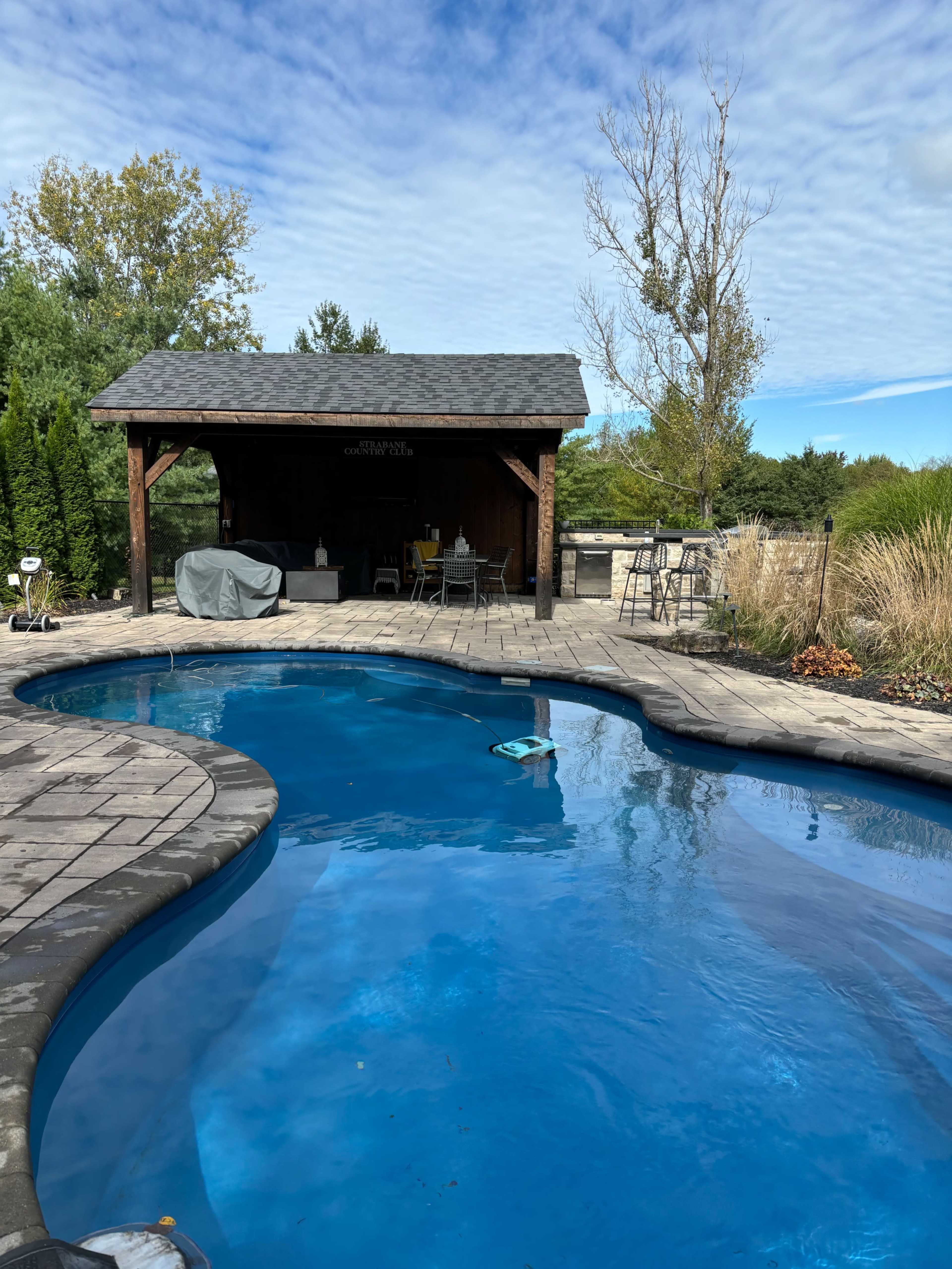 The image depicts a clear swimming pool surrounded by a stone patio, with a wooden gazebo and trees in the background under a cloudy sky.