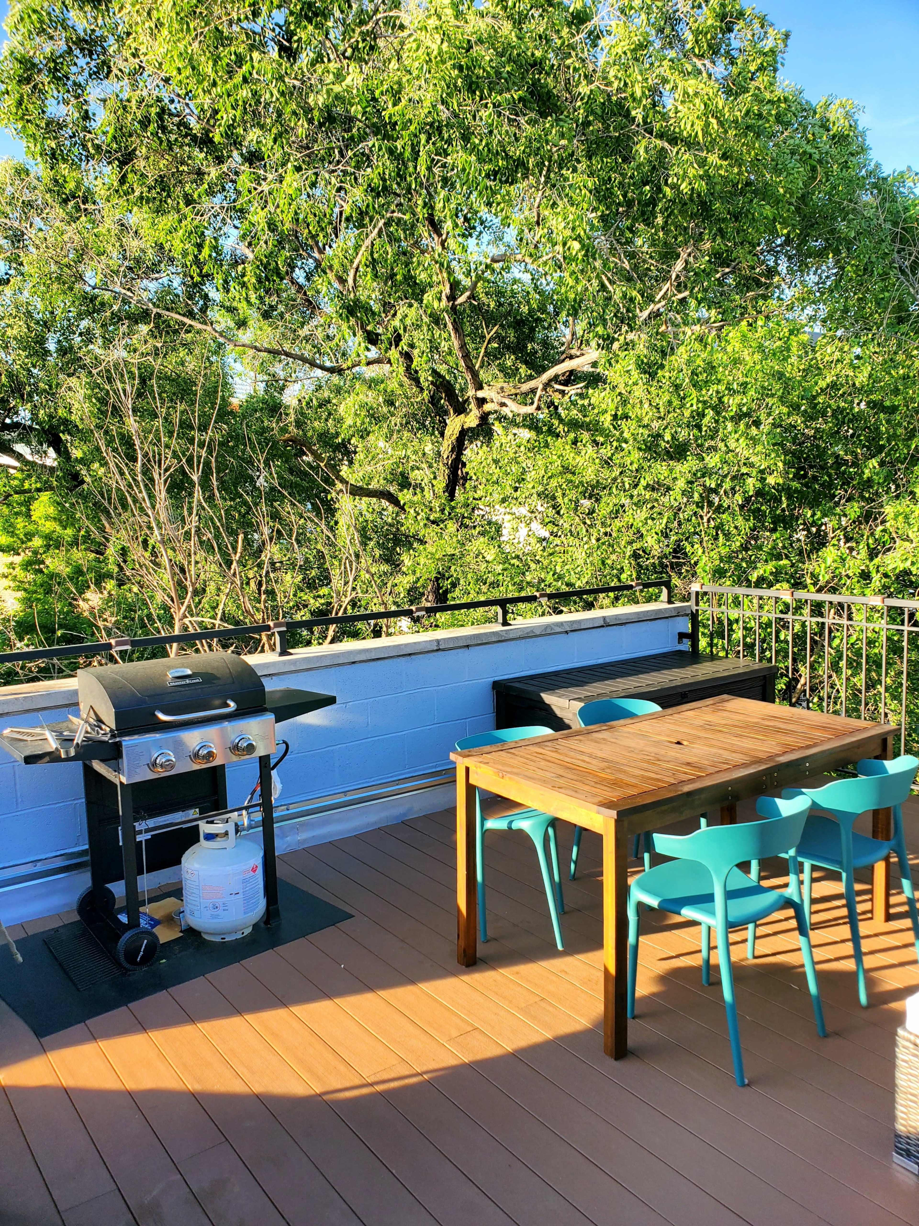 A rooftop deck features a grill and a wooden table surrounded by six blue chairs, with greenery visible in the background.