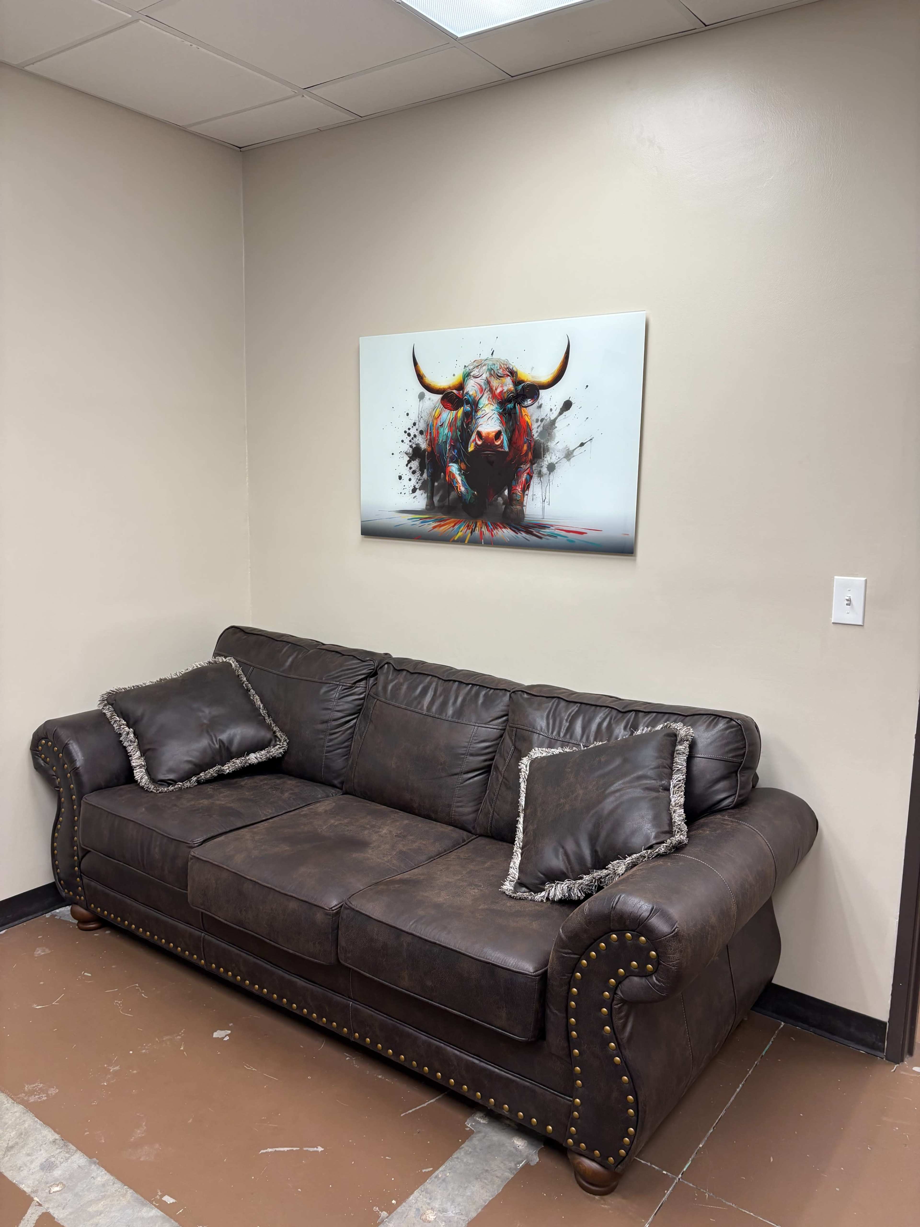 A brown leather couch with decorative pillows is positioned against a beige wall, featuring a colorful bull-themed artwork above it.