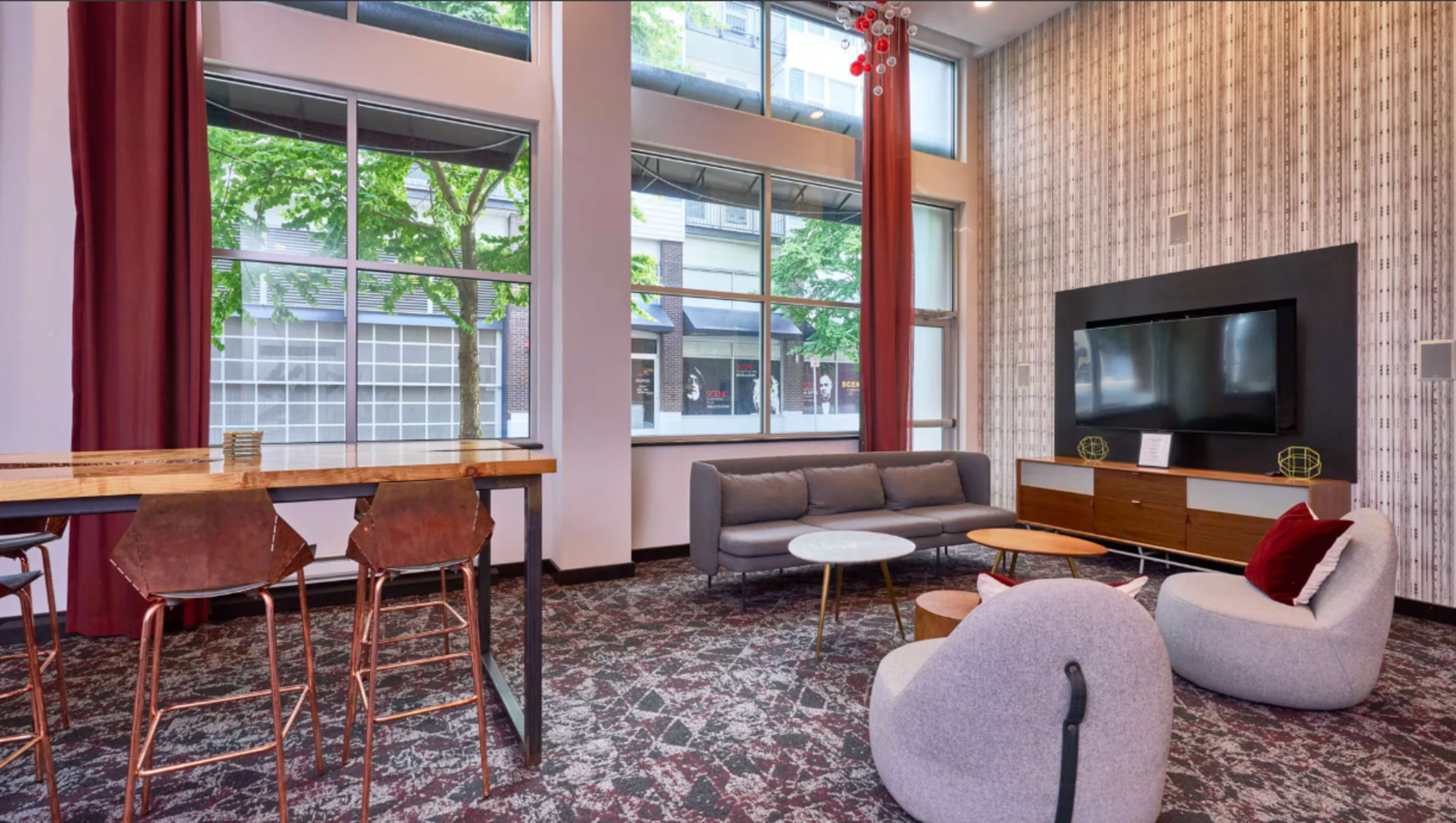 The image shows a modern lounge area featuring a gray sofa, two round tables, a TV unit, and a high bar table with stools, surrounded by large windows letting in natural light.