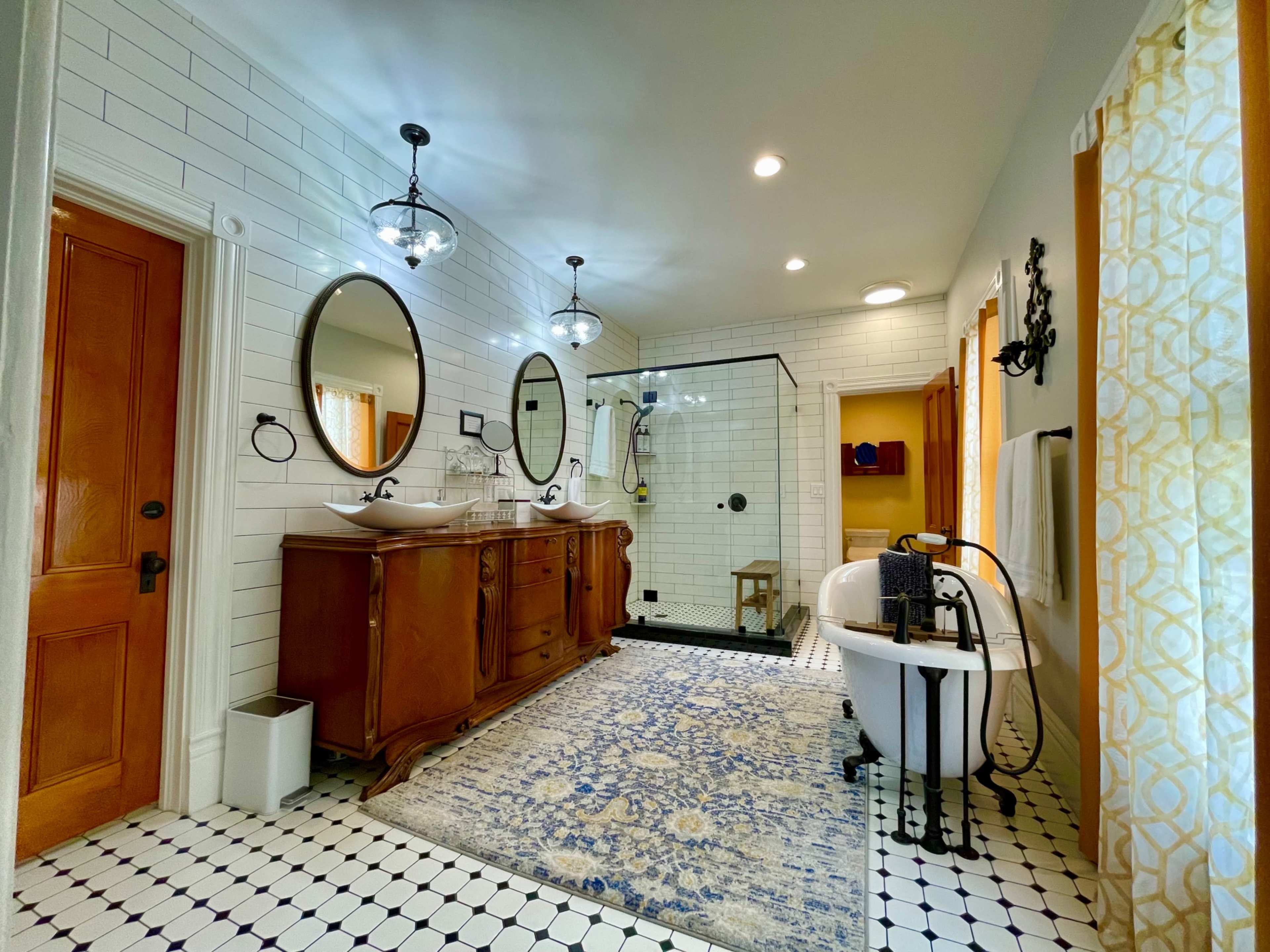A spacious bathroom featuring a wooden double vanity, a freestanding bathtub, and a tiled shower area, all complemented by a patterned rug and decorative lighting.