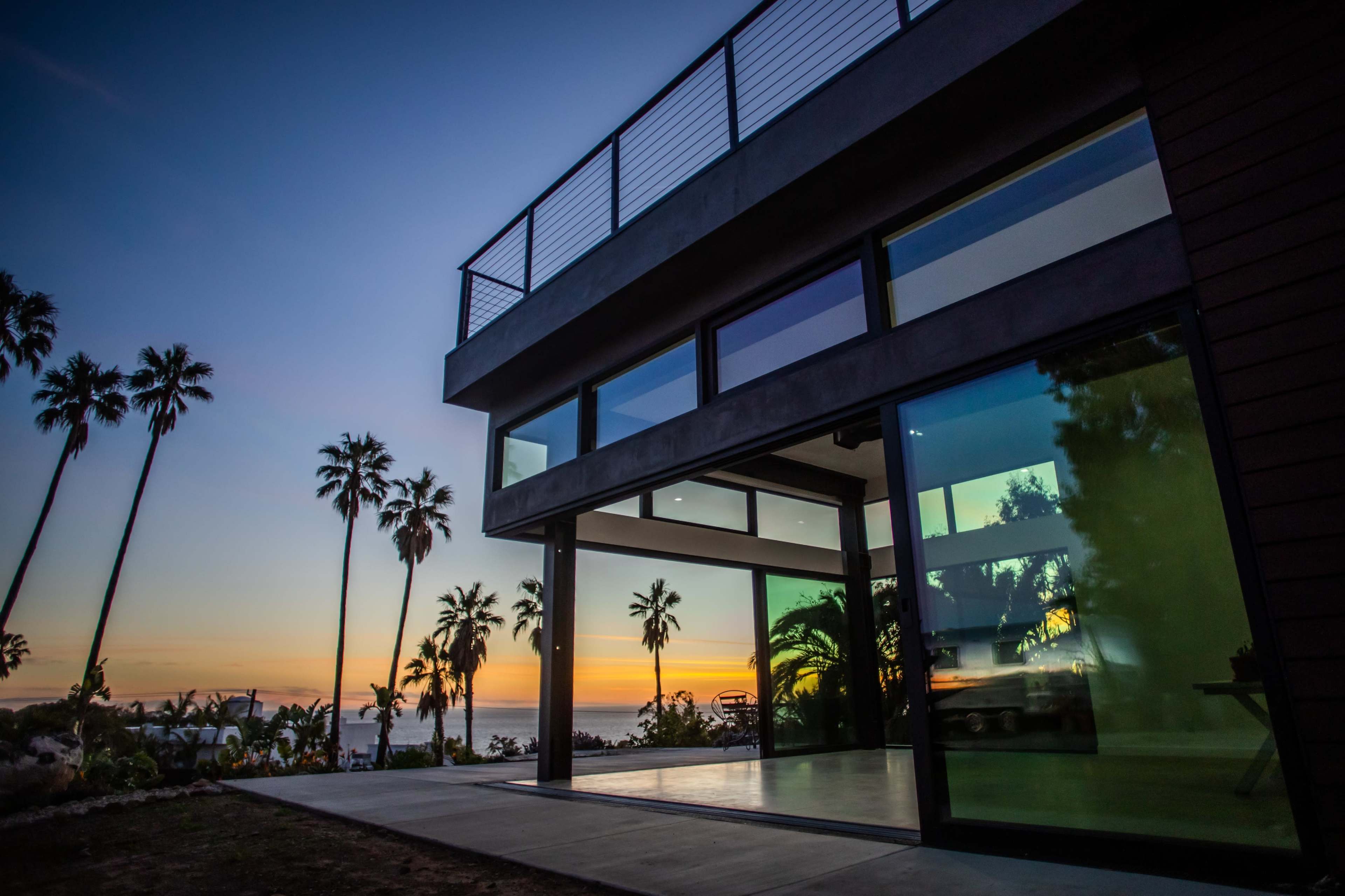 A modern house with large glass windows overlooks a sunset and palm trees.
