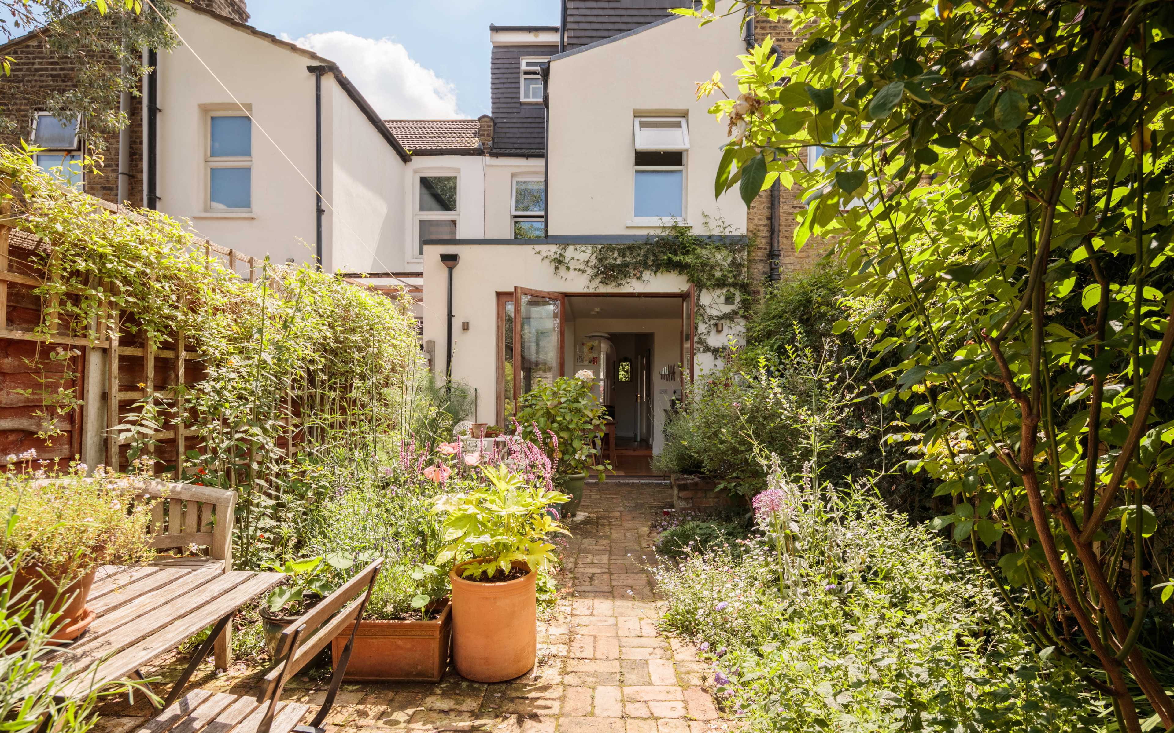 A brick pathway leads to an open doorway surrounded by lush greenery and flowering plants in a residential garden.