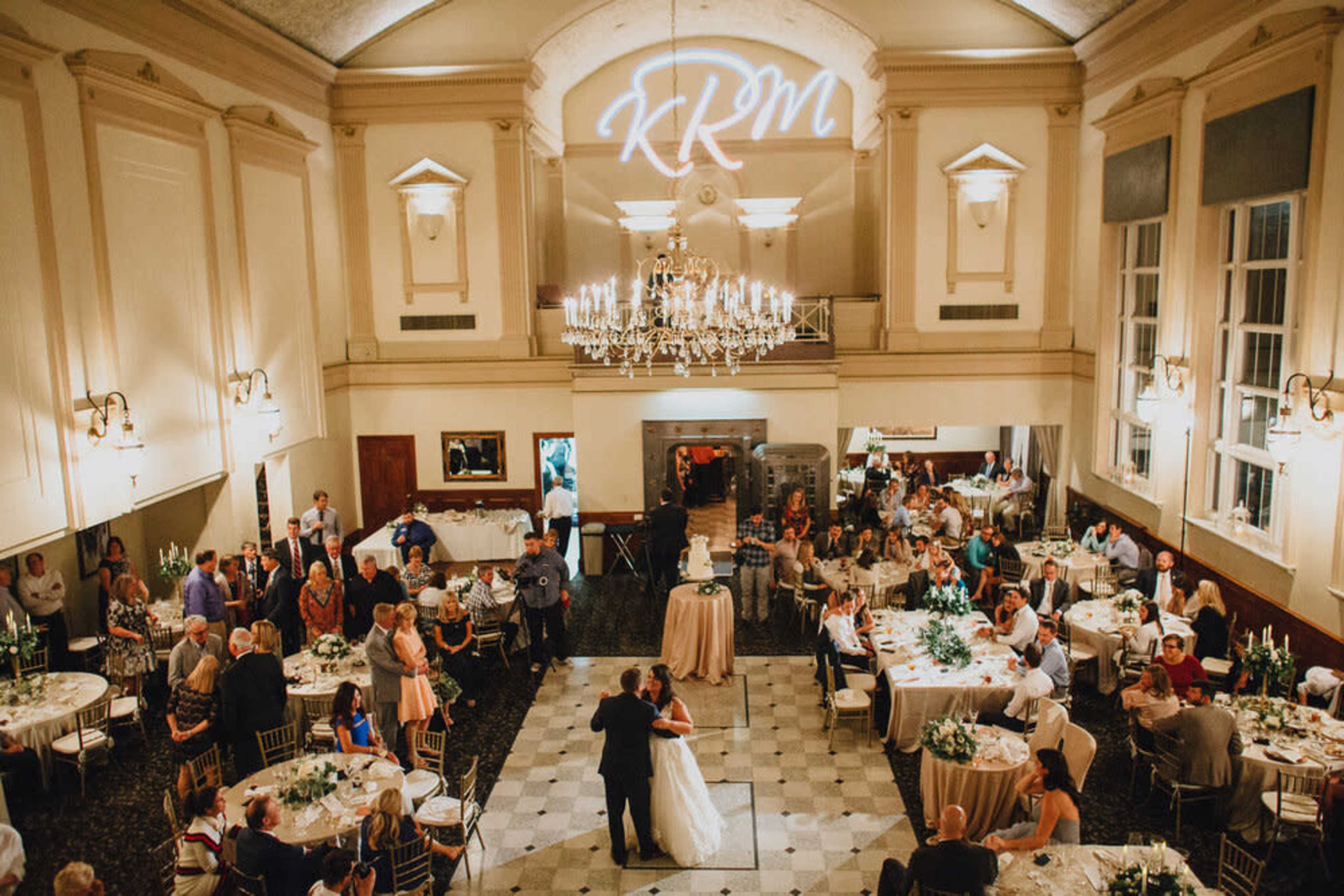 A large banquet hall is filled with guests seated at round tables, while a couple dances at the center beneath a chandelier.