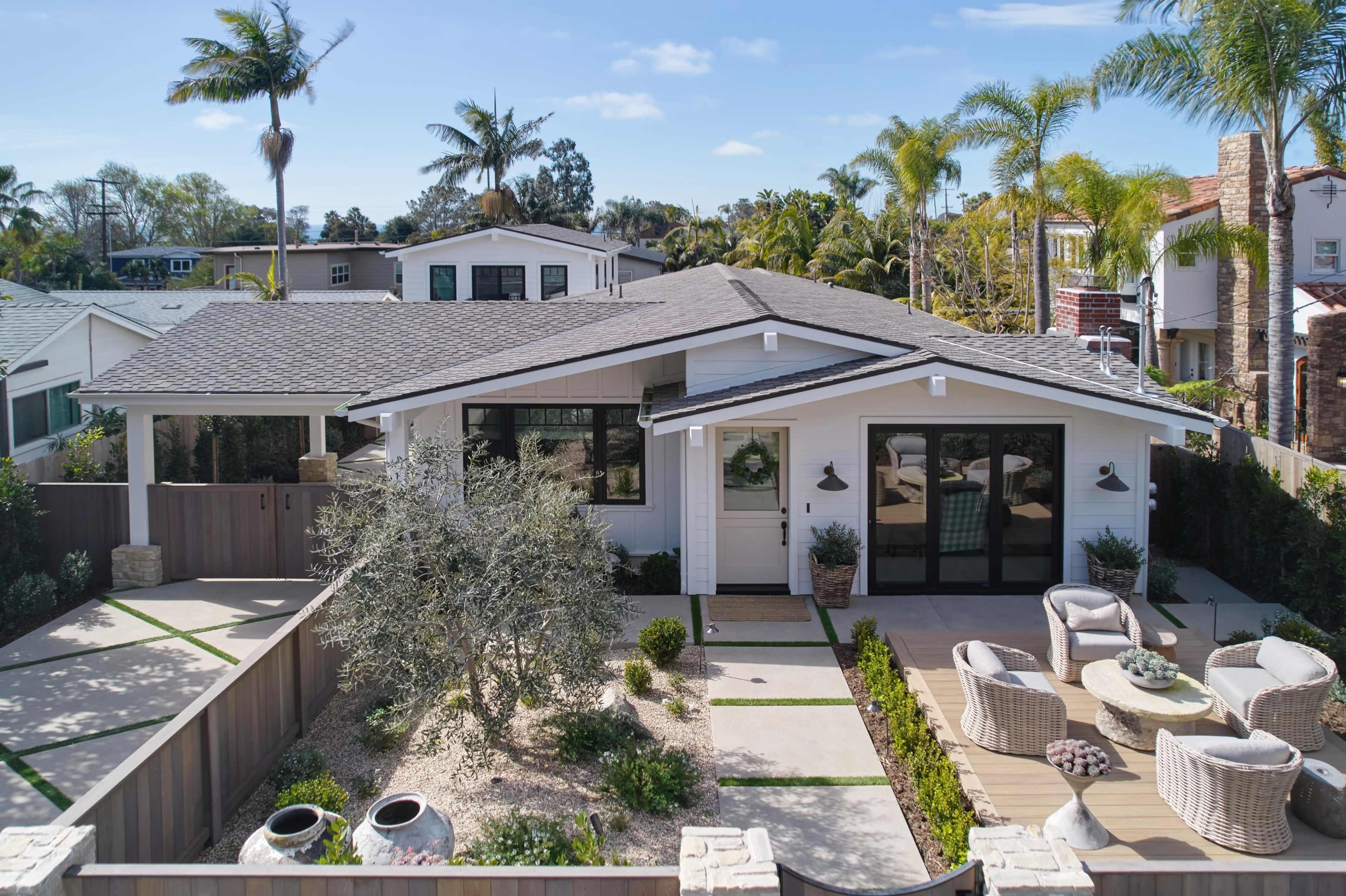 A modern single-story house with a gray roof, surrounded by palm trees and a landscaped yard featuring a patio area with outdoor furniture.