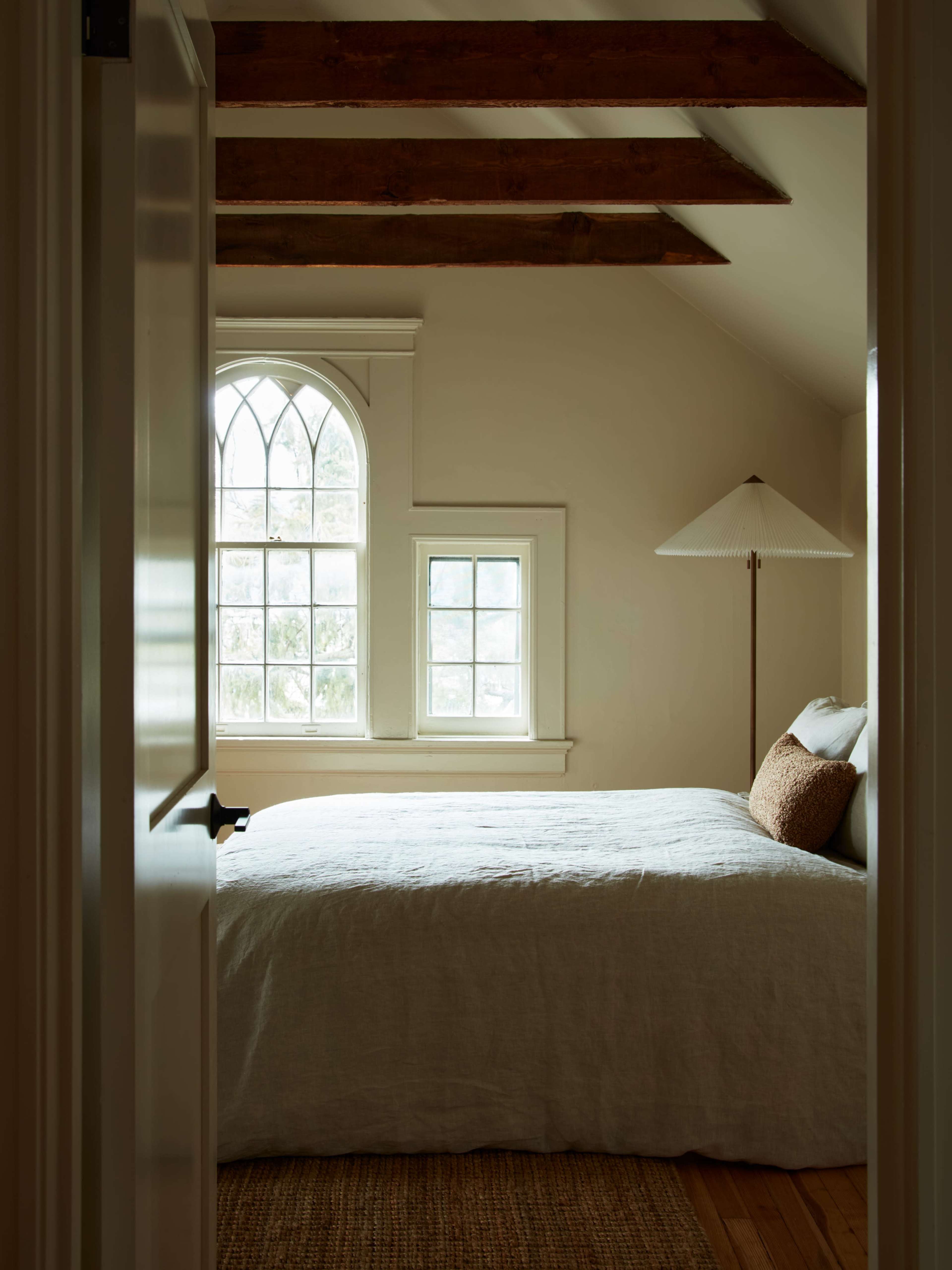 A neatly arranged bedroom features a bed with a light-colored quilt, a decorative pillow, and a floor lamp beside a window with arched framing.