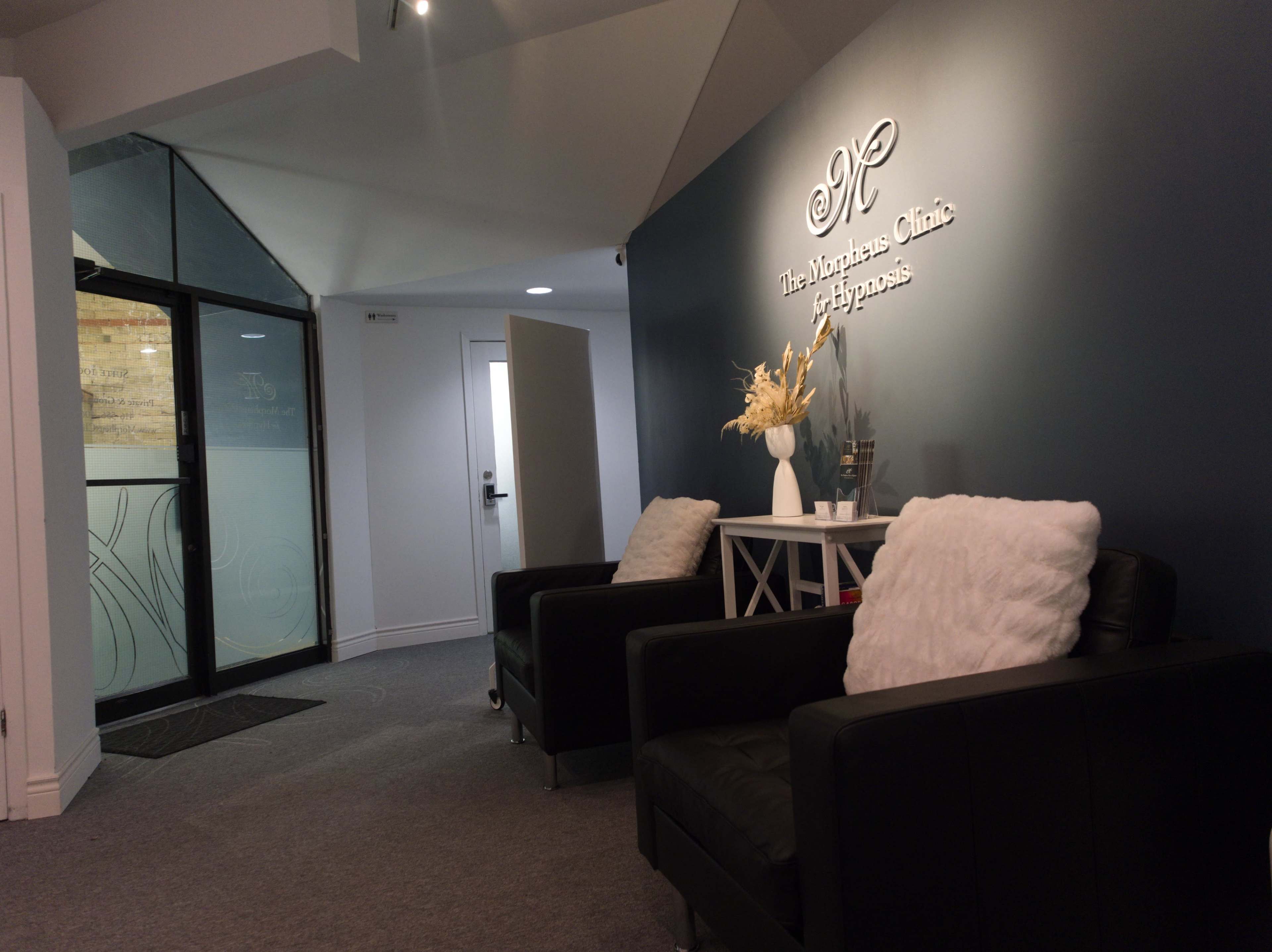 The image shows a modern reception area with two black chairs, a small table, and a sign on the wall displaying the clinic's name.