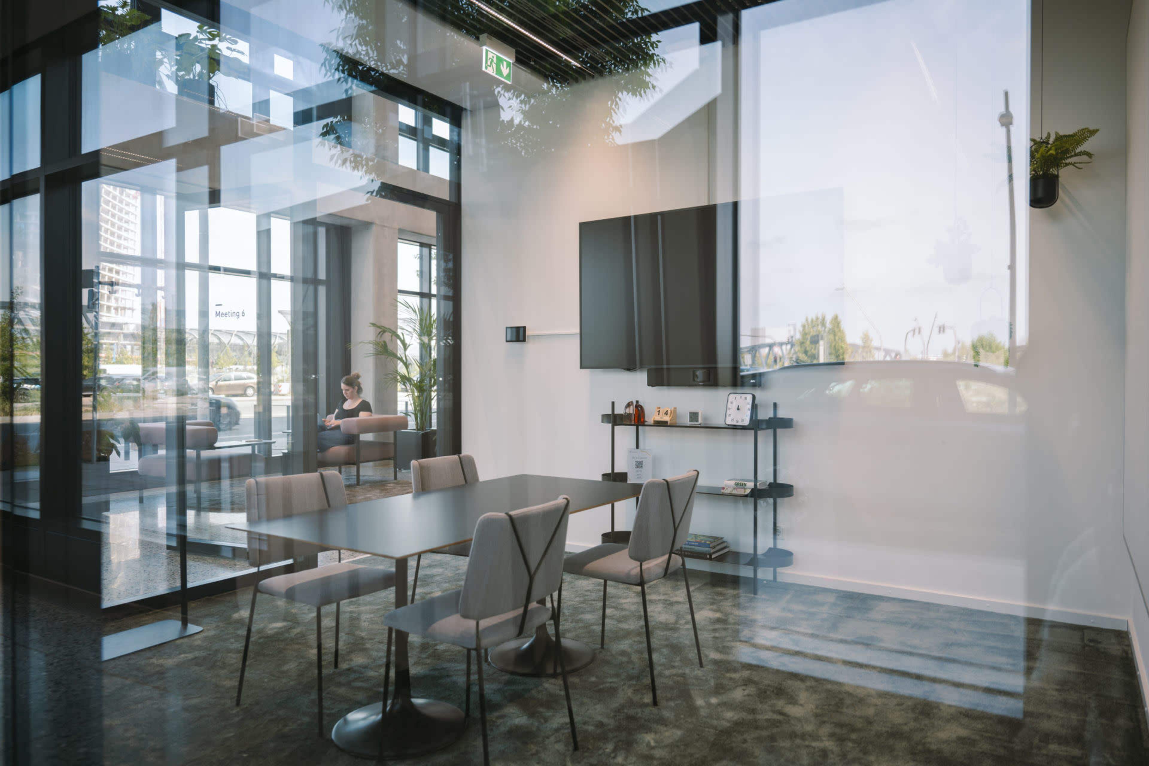 A modern conference room features a large table surrounded by chairs, with a television mounted on the wall and large windows providing a view of the exterior.