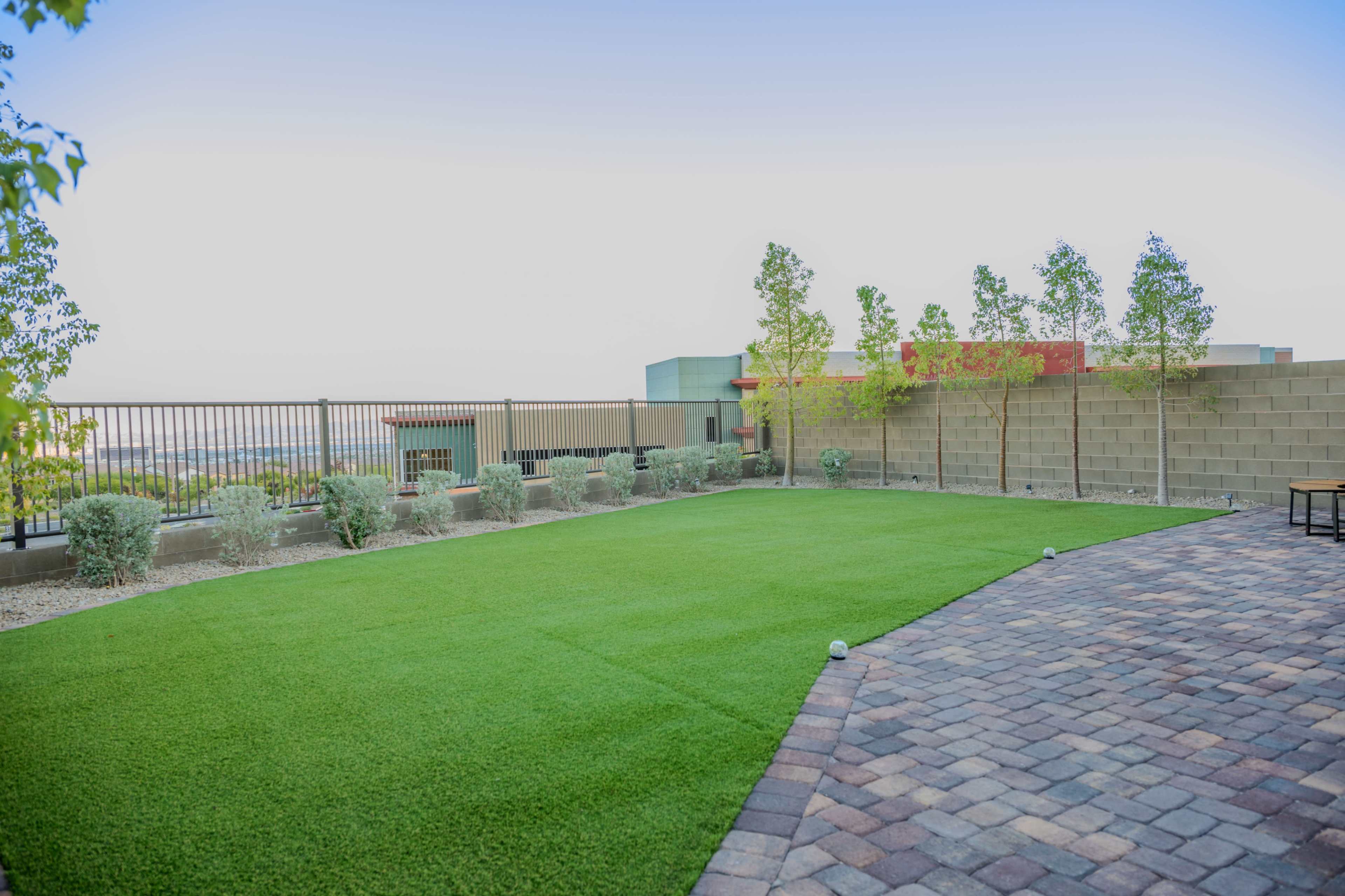The image shows a landscaped outdoor area featuring a green lawn and a brick paved patio, surrounded by low shrubs and a fence.