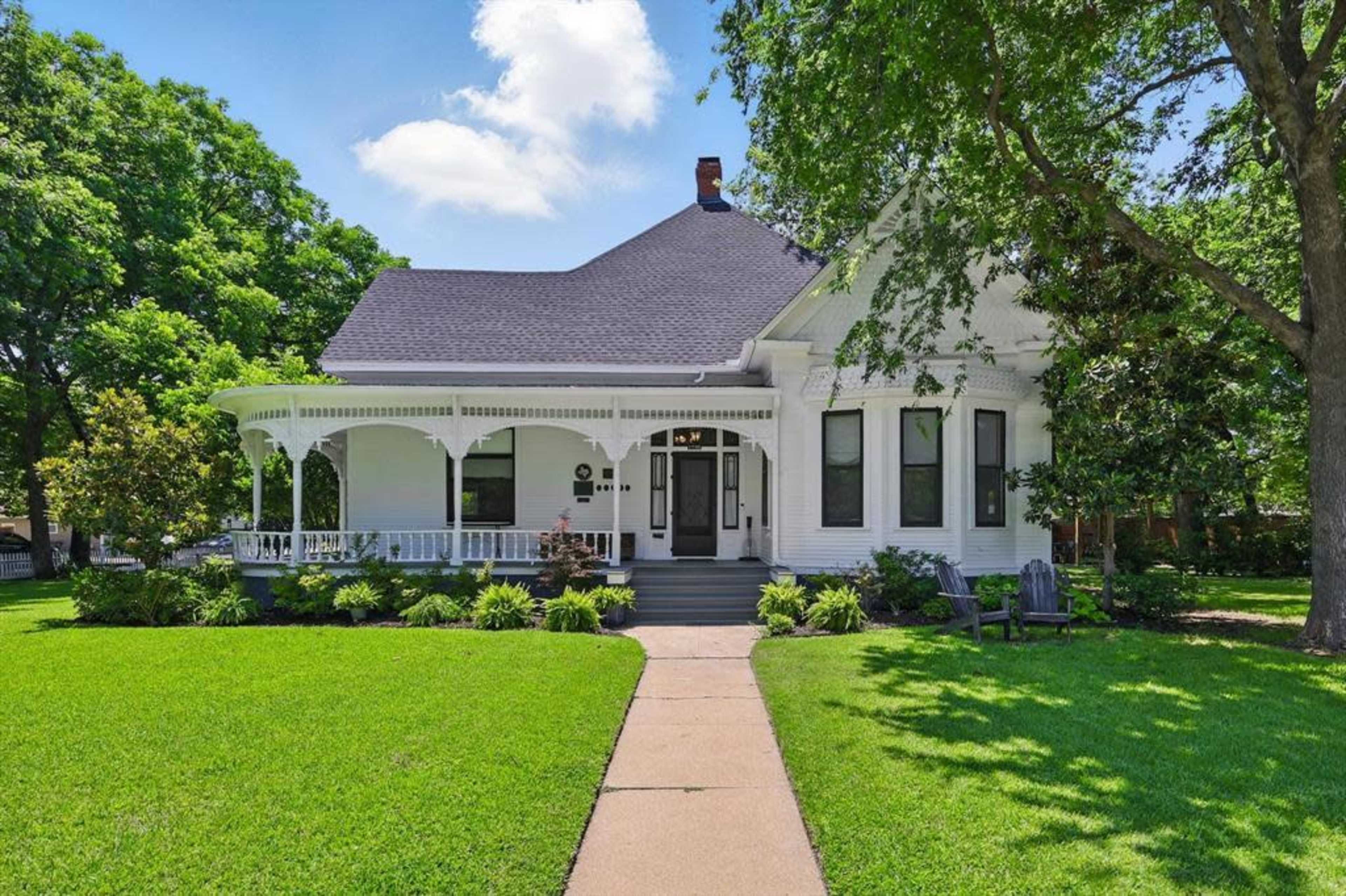 The image shows a large, white Victorian-style house with a wraparound porch, surrounded by a well-maintained lawn and mature trees.