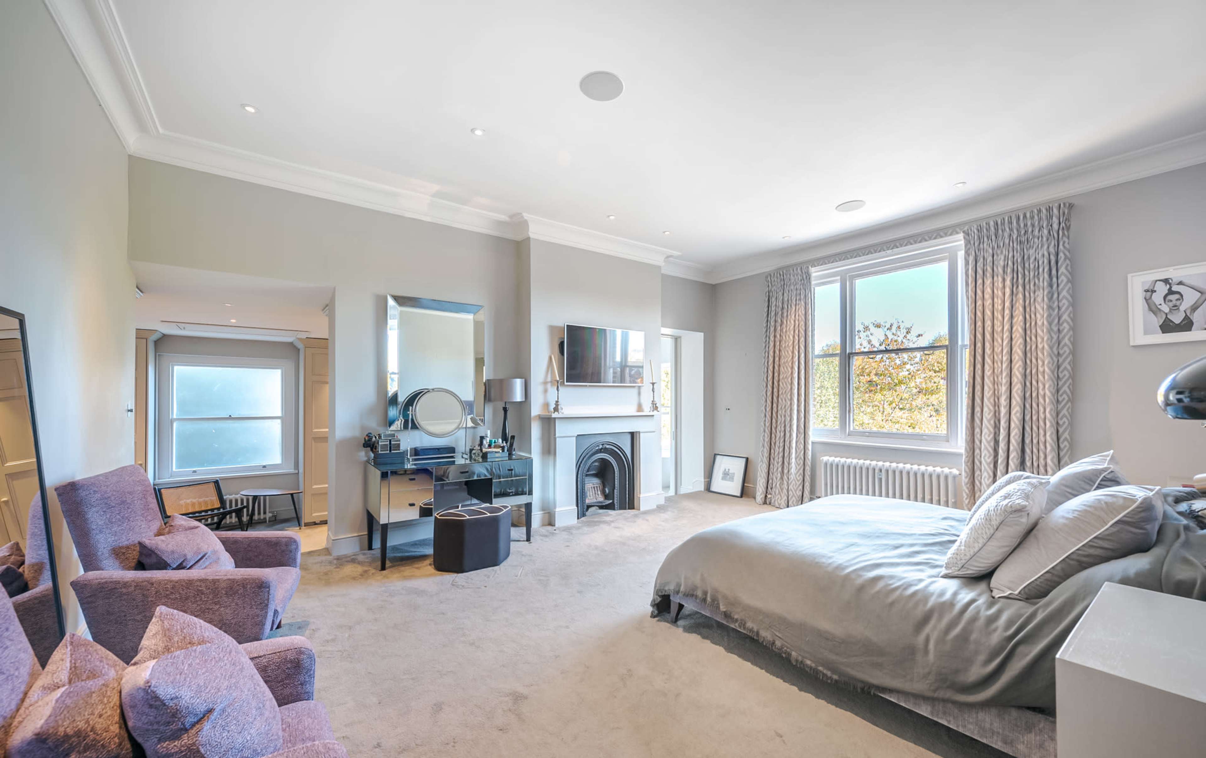 The image shows a spacious bedroom with a large bed, a fireplace, a mirror-desk setup, and two purple armchairs.