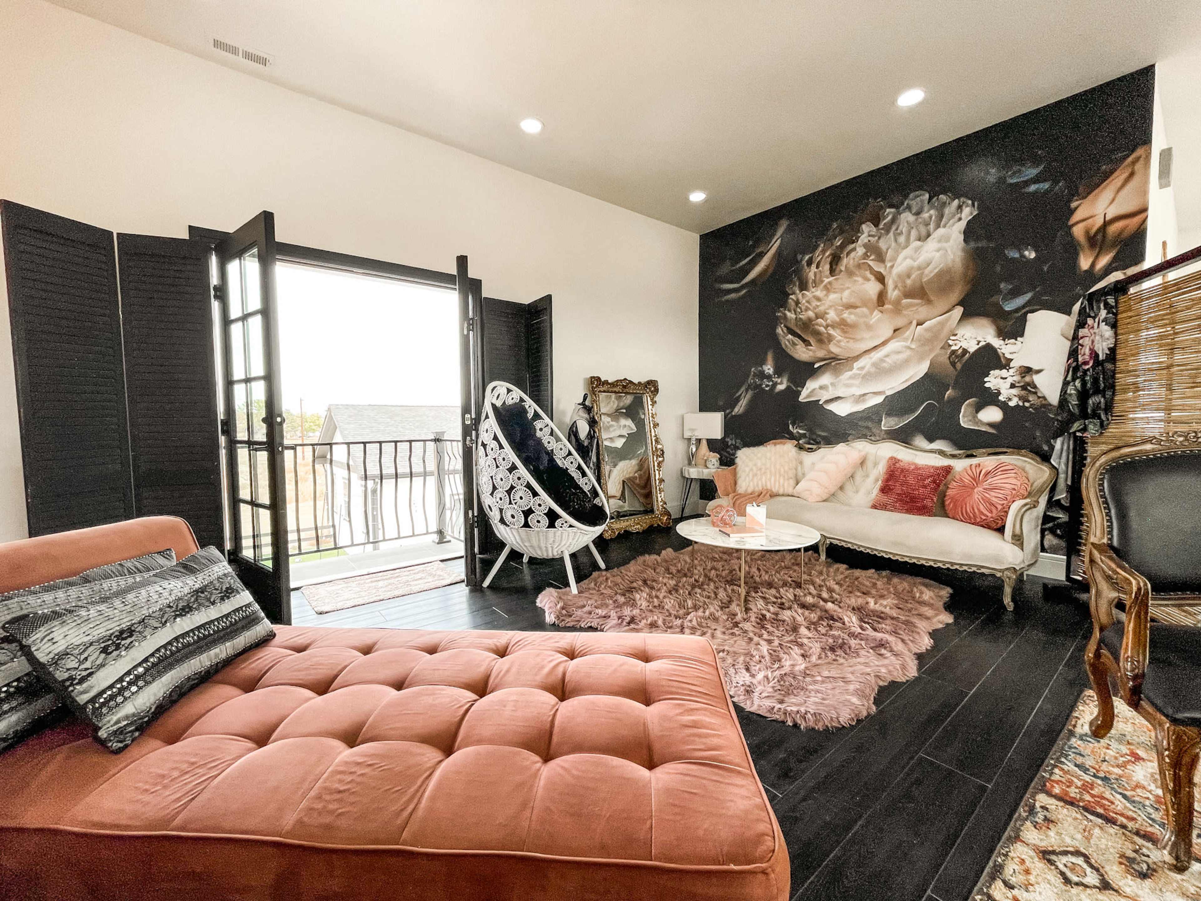 A stylish living room with a pink velvet sofa, a white hanging chair, and large windows opening to a balcony, accented by a floral mural on one wall.