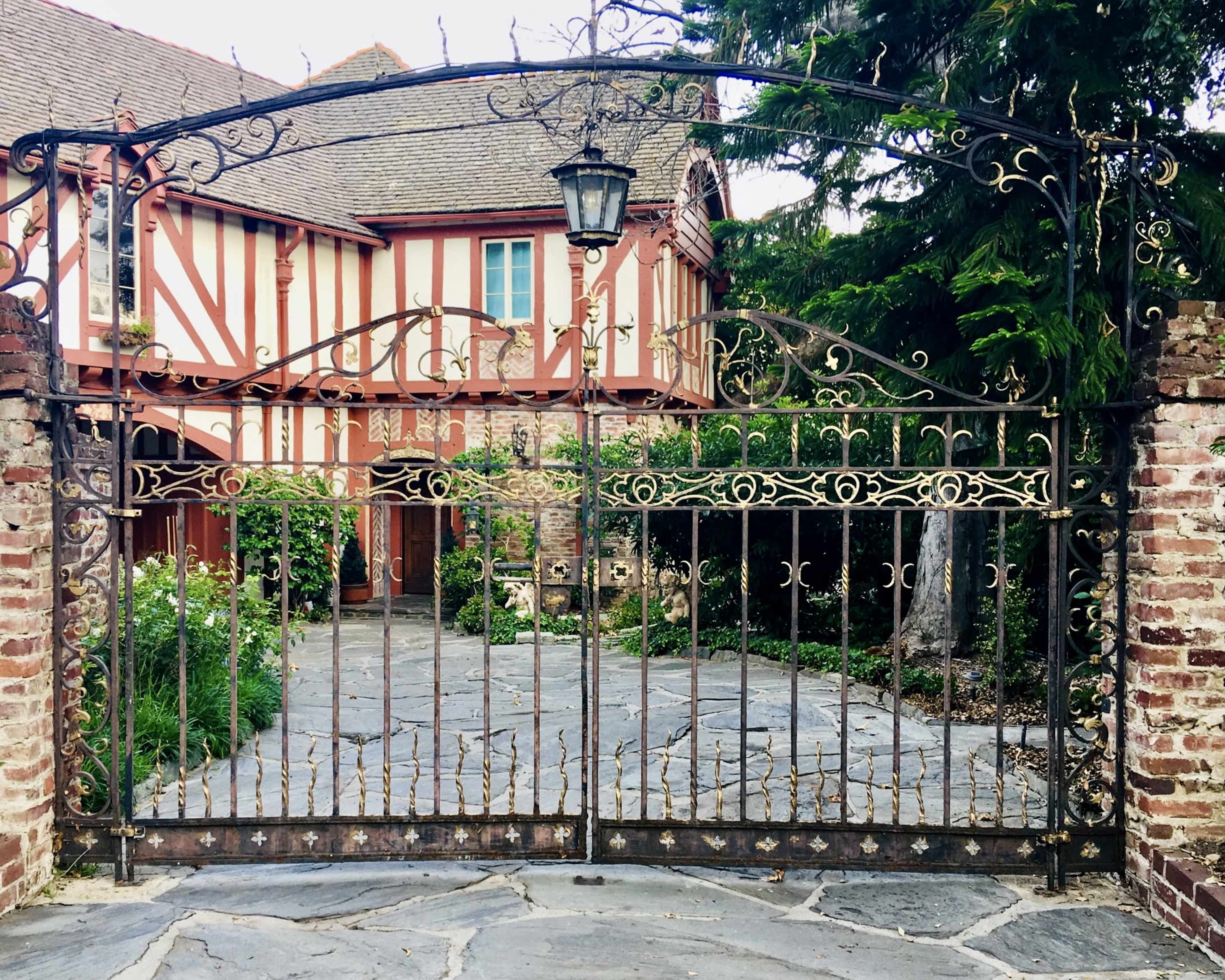 A wrought iron gate with intricate designs encloses a stone pathway leading to a red and white striped house surrounded by greenery.