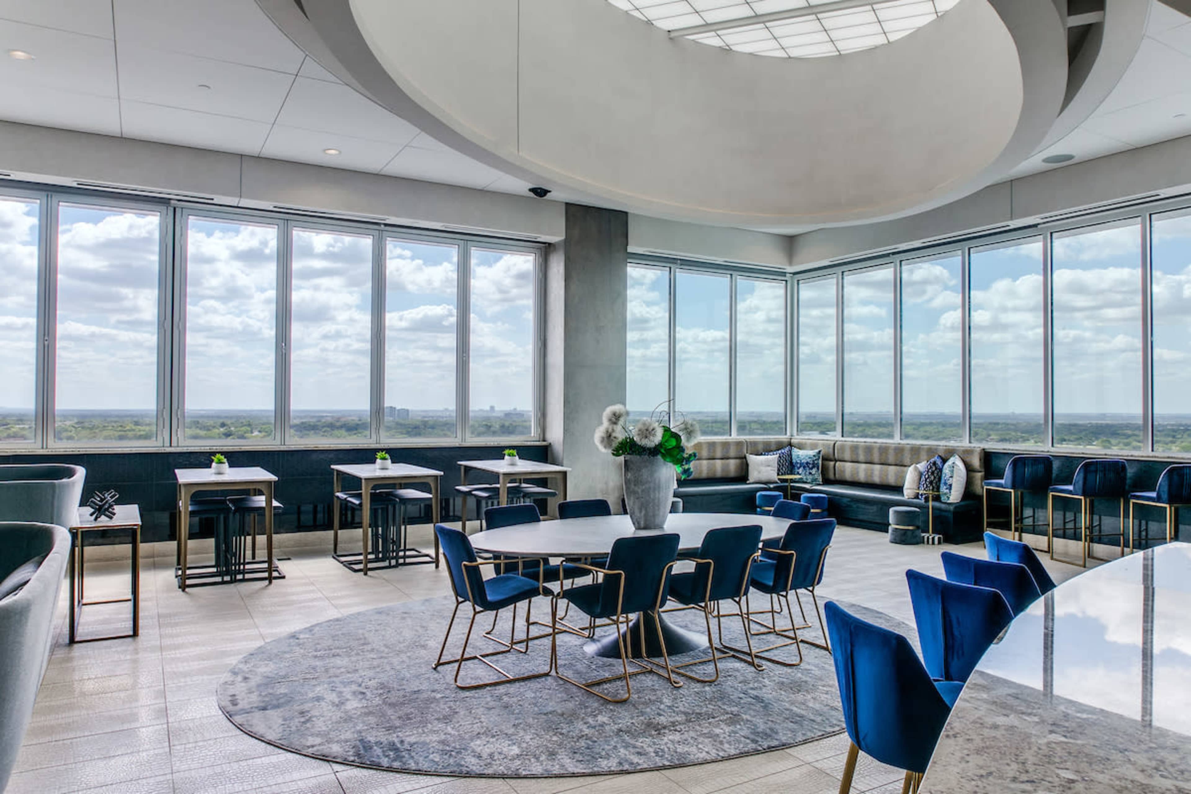 A modern interior features a circular space with large windows offering a view, a round table surrounded by blue chairs, and a lounge area with low seating.